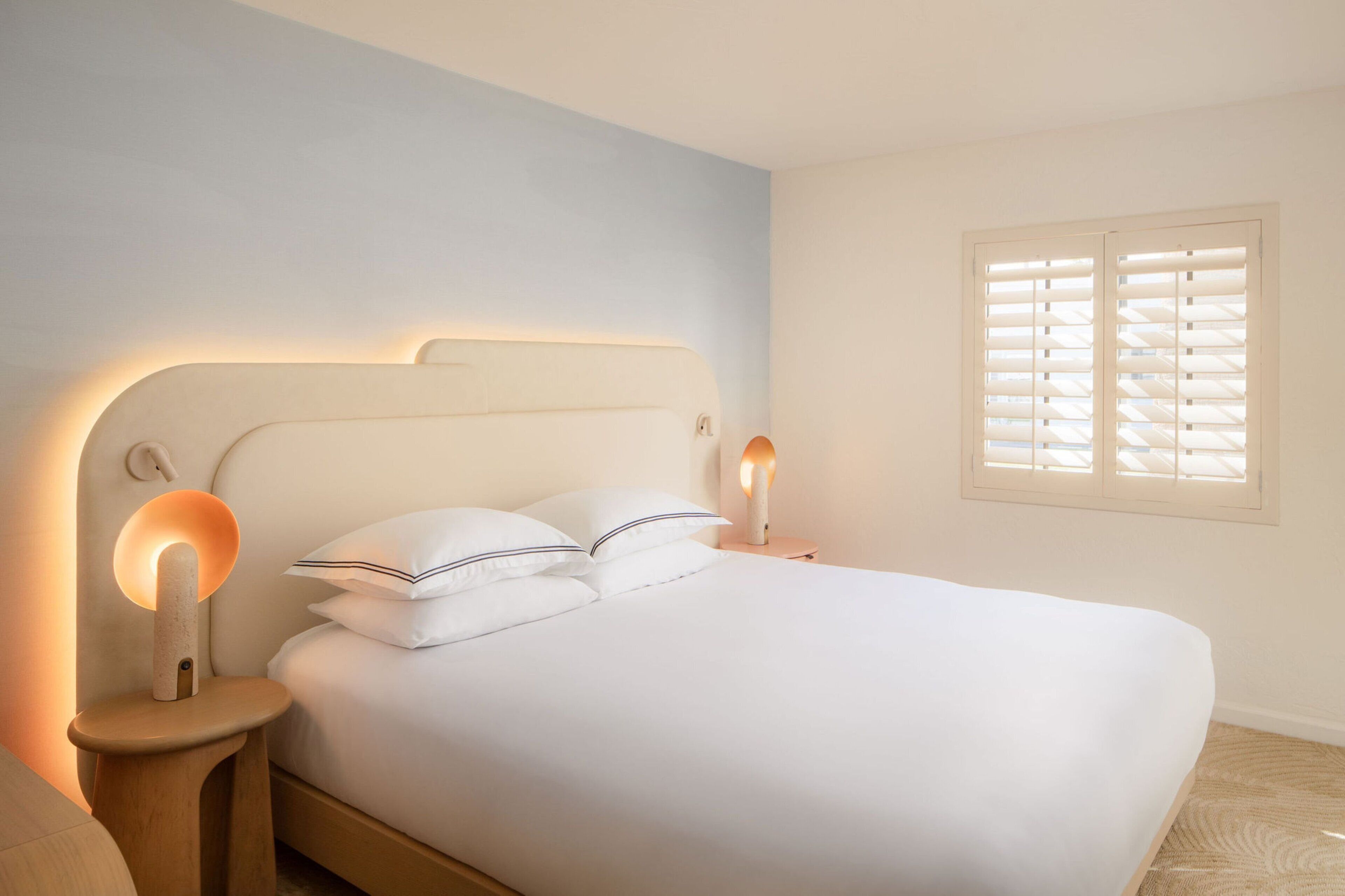 A neatly made bed with two pillows, a bedside table with a lamp, and a window with blinds.