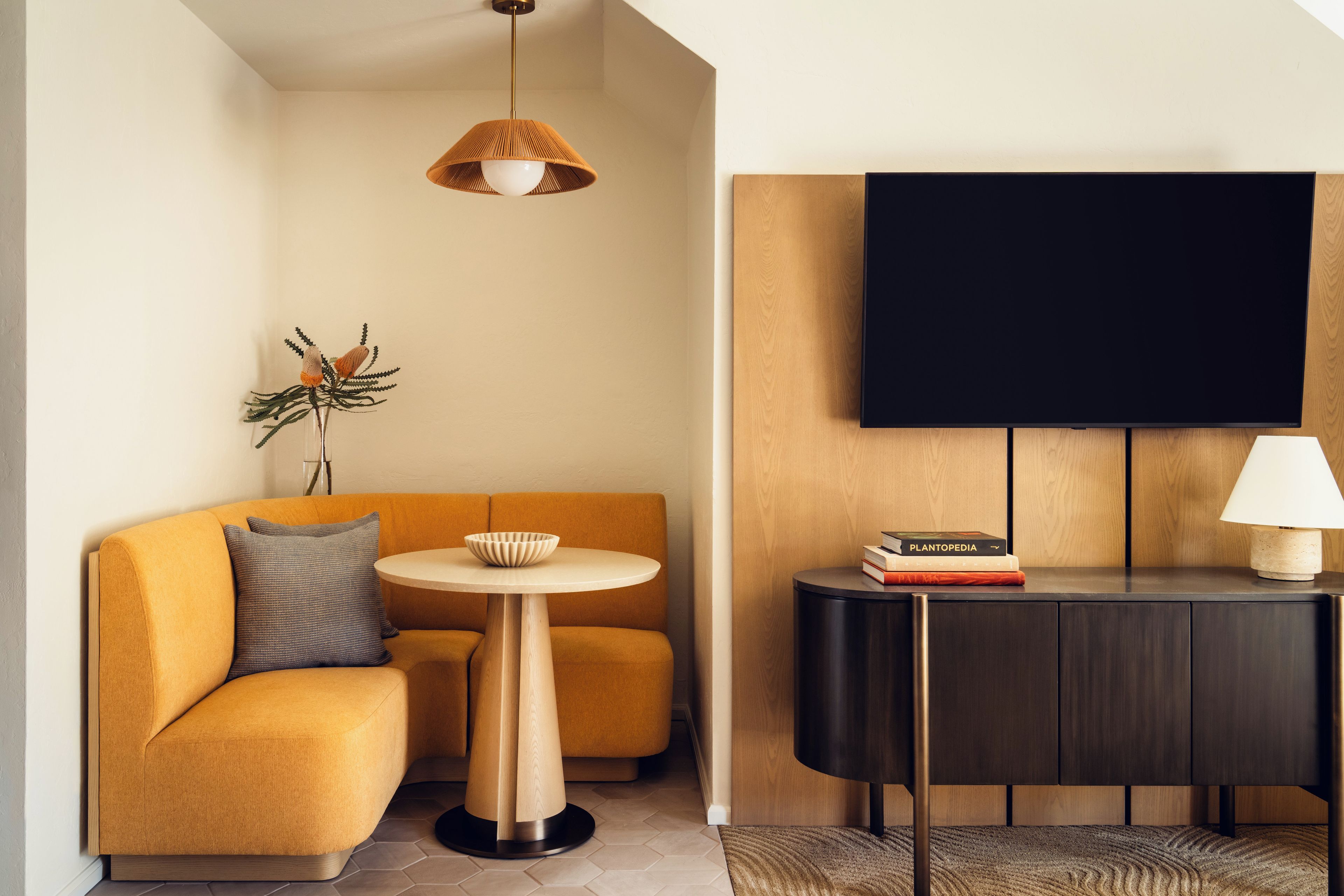 A modern living room with a sofa, a small round table, a flat-screen TV mounted on the wall, and a side table with books and a lamp.