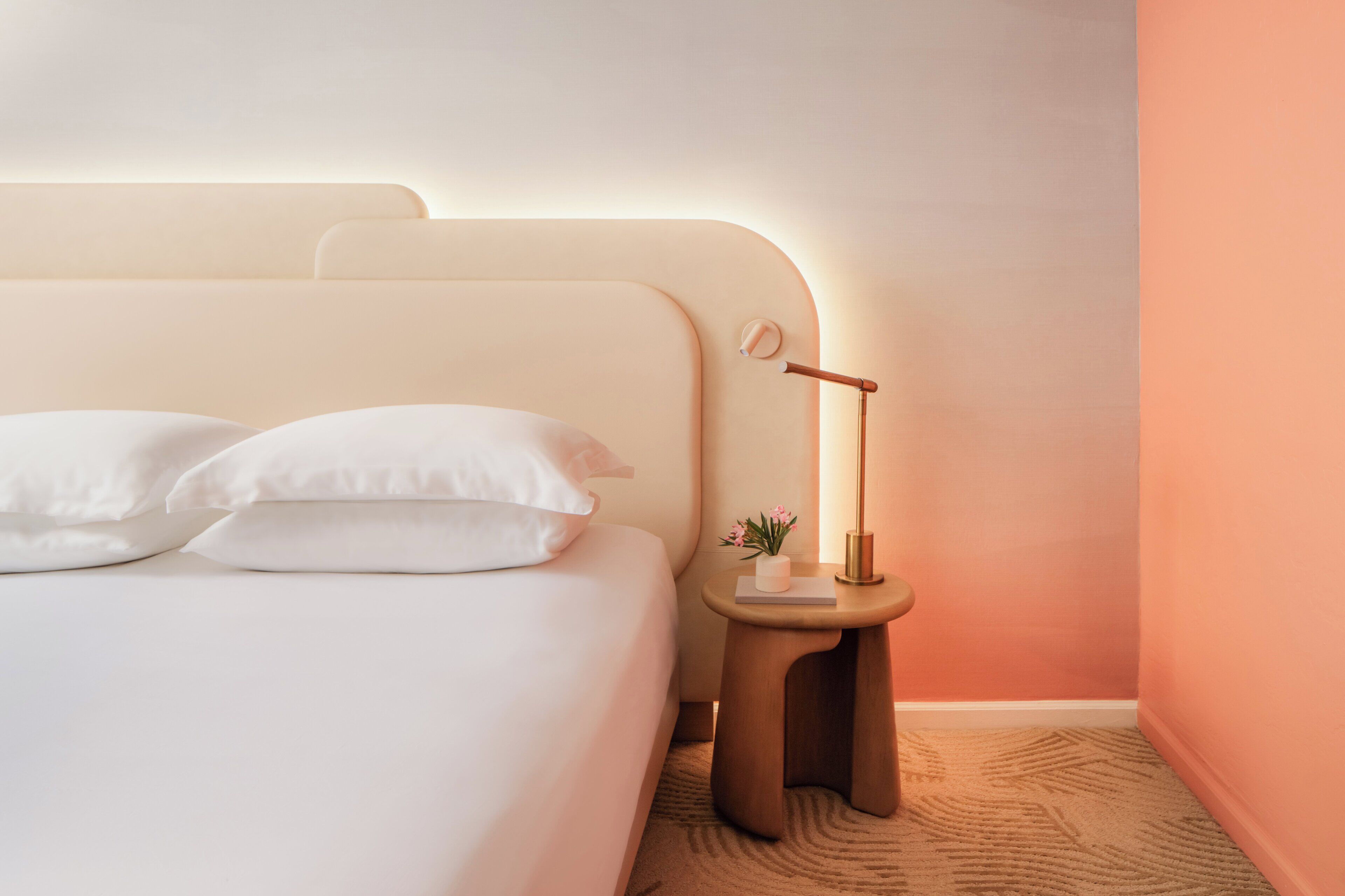 A neatly made bed with white linens and pillows, a bedside table with a lamp, and a small plant.