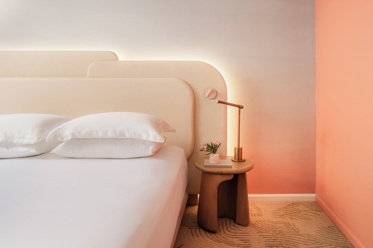 Ein ordentlich bezogenes Bett mit weißen Leinen und Kissen, ein Nachttisch mit einer Lampe und eine kleine Pflanze.