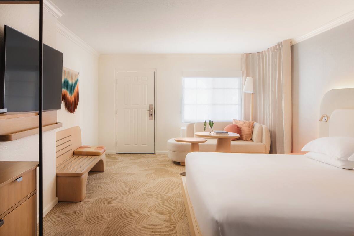Habitación de hotel con cama, sofá, mesita, banca, televisor y puerta.