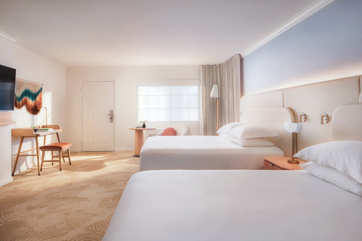 Ein Hotelzimmer mit zwei Betten, einem Schreibtisch, einem Stuhl, einer Lampe und einem großen Fenster.