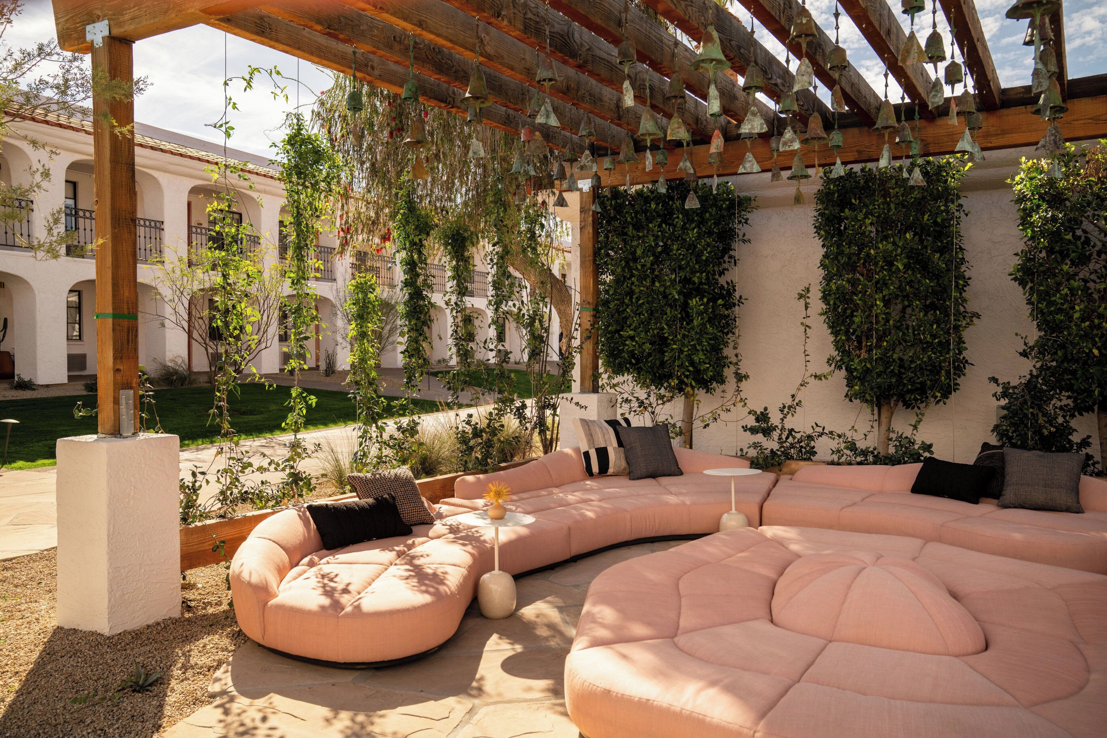 A covered outdoor seating area with a curved sofa and hanging bells, surrounded by greenery and a building with arches in the background.