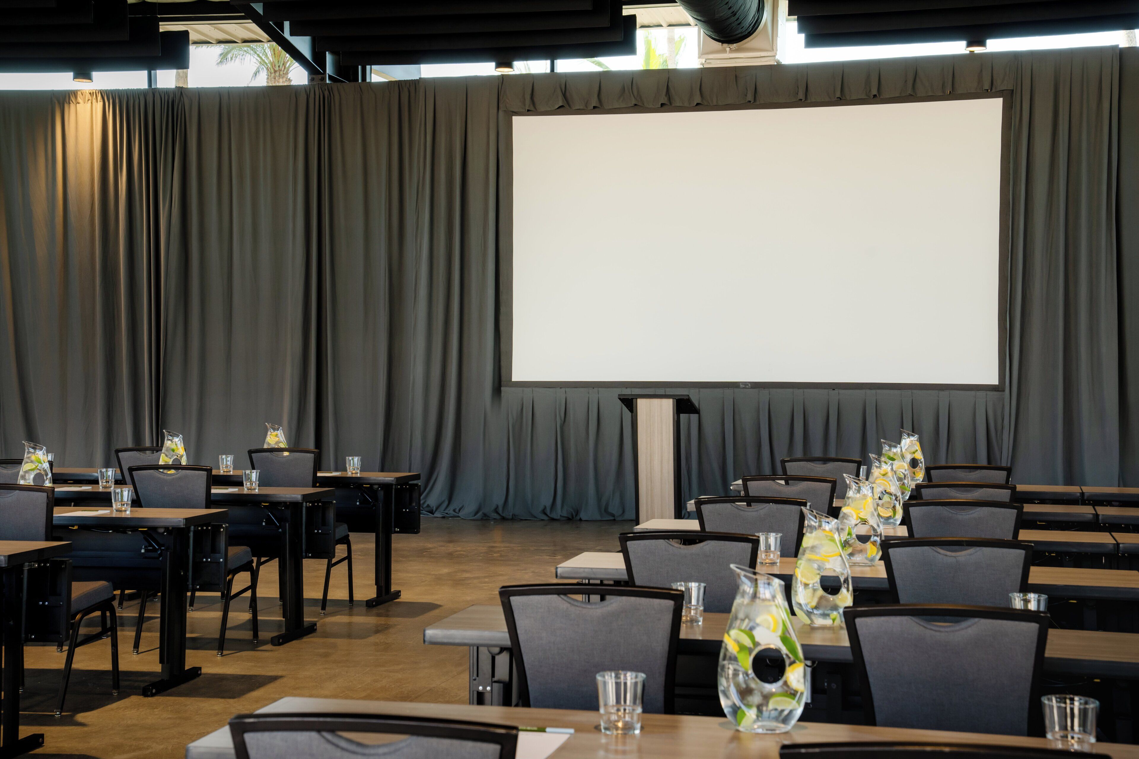 Un salón de conferencias con una pantalla grande, mesas, sillas y jarrones decorativos.
