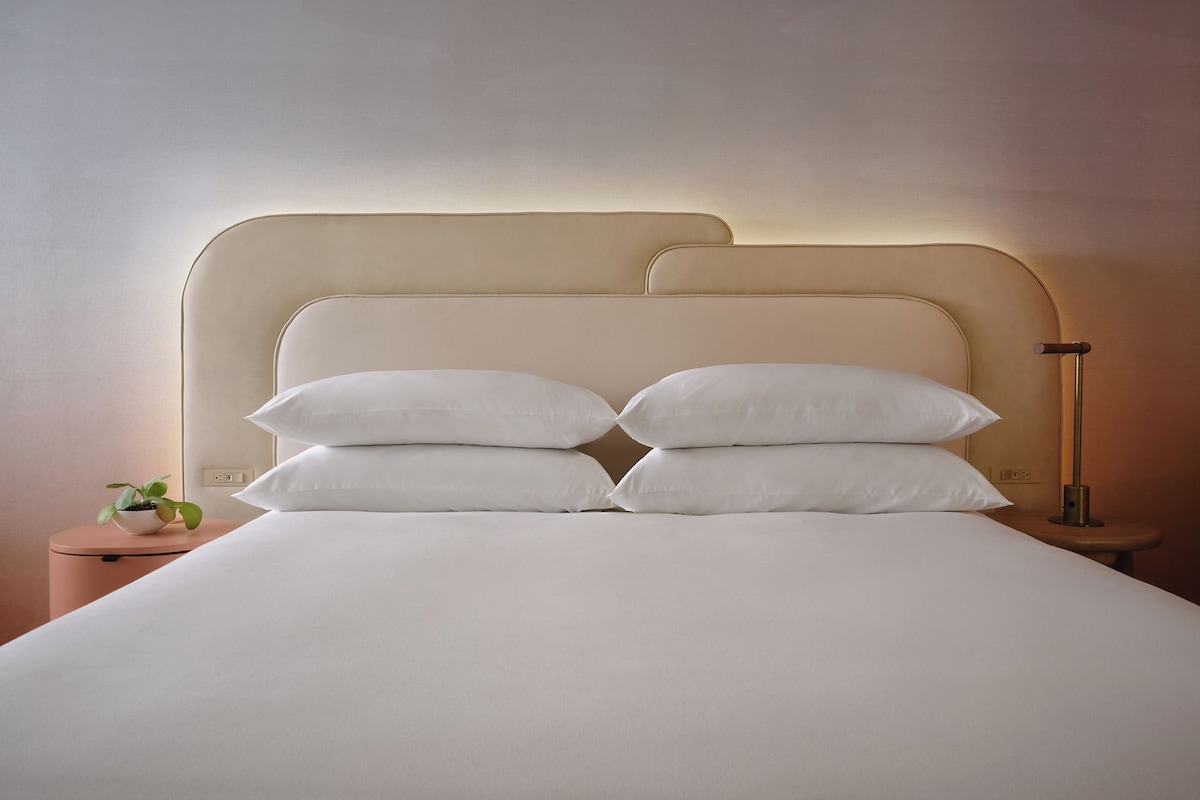 A neatly made bed with white linens and pillows, a bedside plant, and a modern lamp on a bedside table.