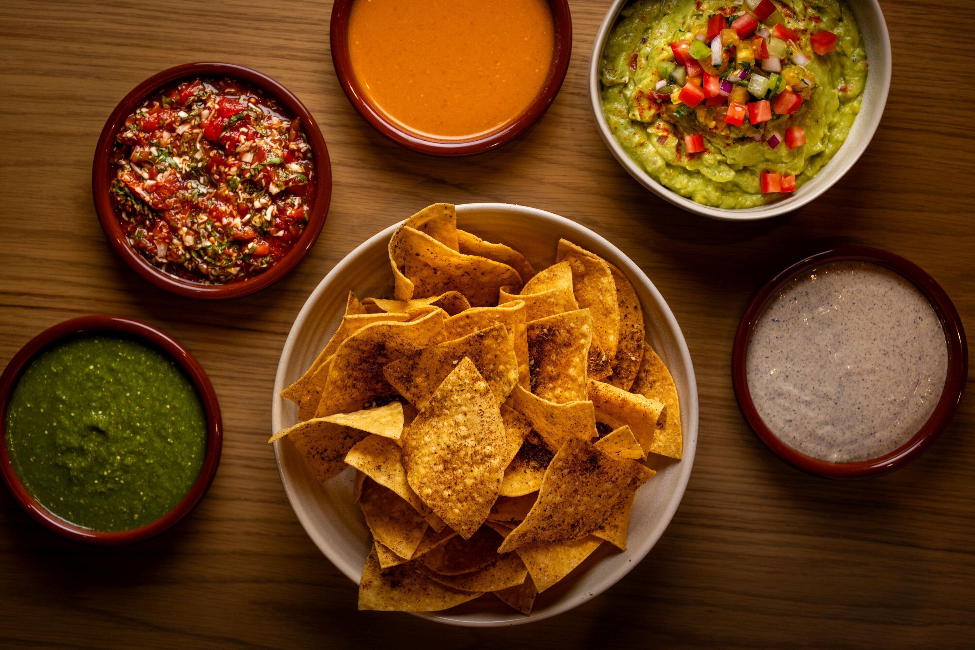 A bowl of nachos with various dips on a wooden table.