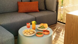 Un sofá gris con un almohadón naranja, una mesa redonda con una bebida, un cuenco de patatas fritas, un bol de ensalada, un panecillo y dos recipientes pequeños de salsas.