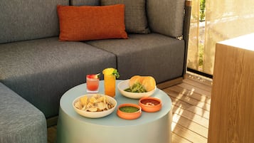 A grey sofa with an orange pillow, a round table with a drink, a bowl of chips, a bowl of salad, a bread roll, and two small bowls of sauces.
