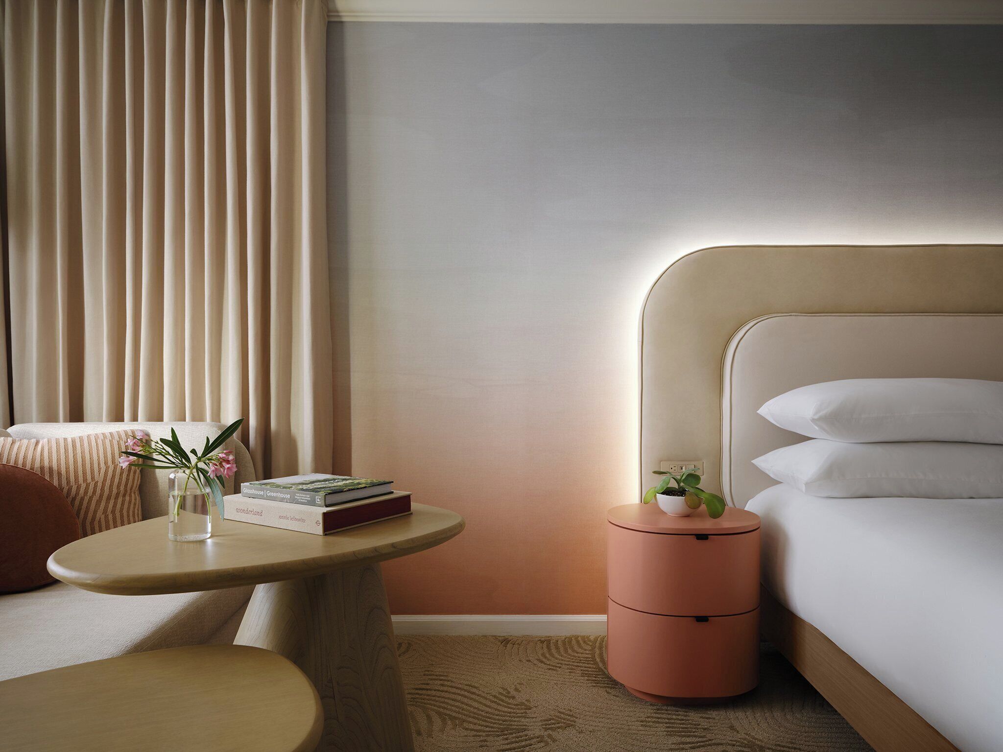 Una habitación de hotel con una cama, una mesita de noche con libros y un jarrón, una mesa redonda con una silla y una pequeña planta en maceta.