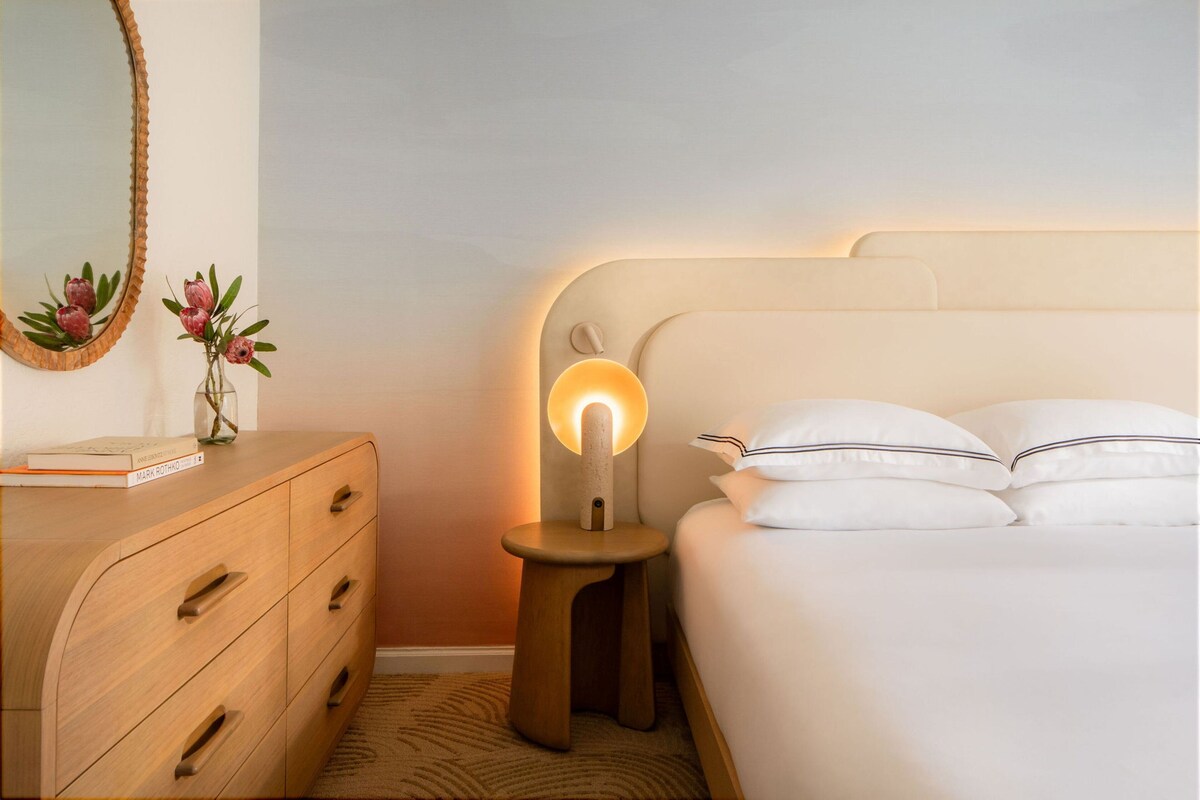 Ein ordentlich eingerichtetes Schlafzimmer mit einem hölzernen Schminktisch, einer Nachttischlampe und einem Bett mit weißer Bettwäsche.
