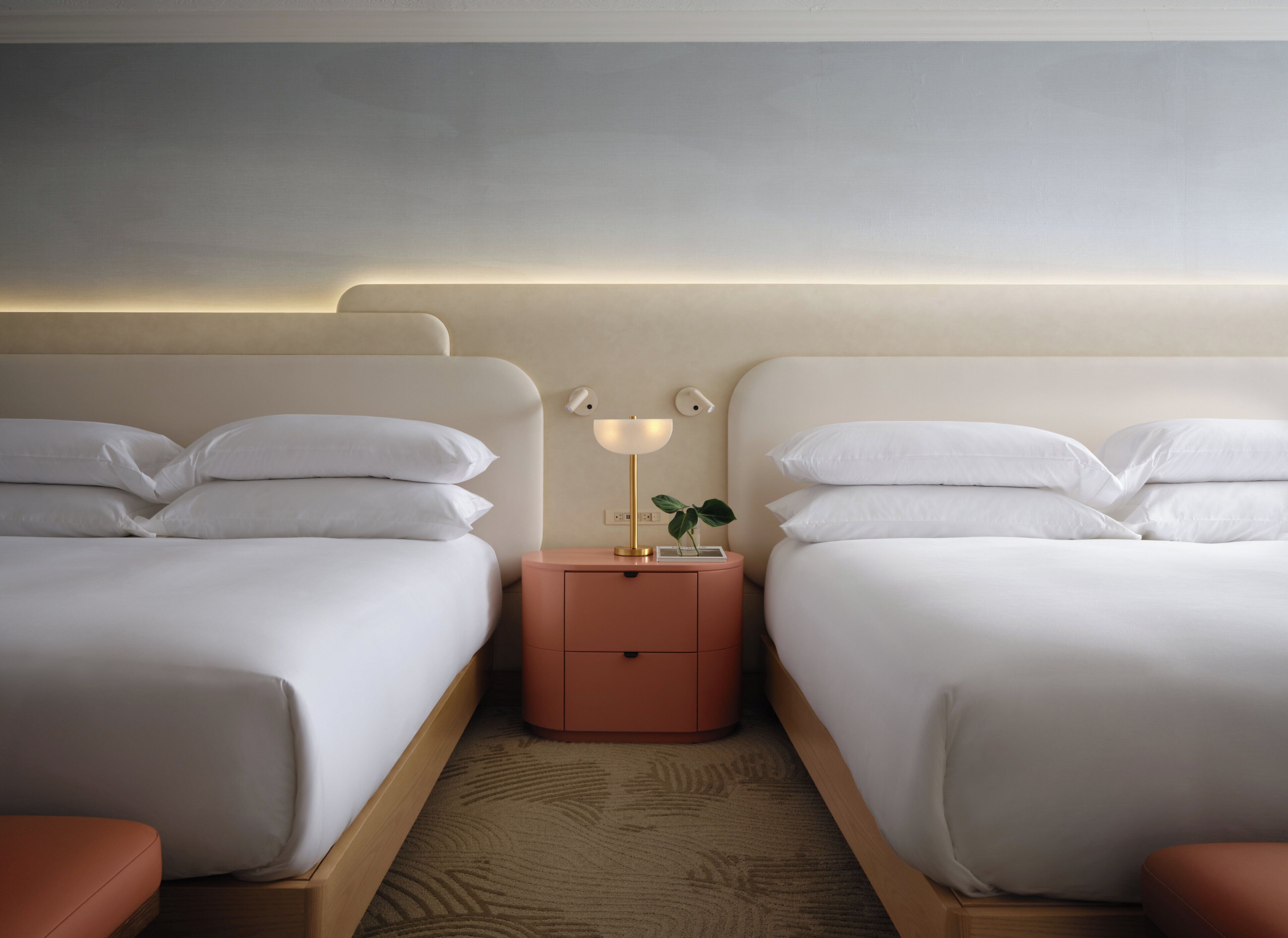 Quarto de hotel com duas camas, uma mesa de cabeceira com abajur e uma pequena planta.