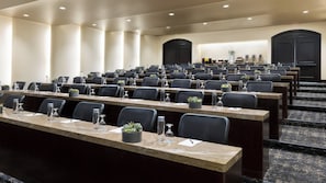 Un salón de conferencias con filas de mesas y sillas, equipado con botellas de agua y pequeñas macetas.