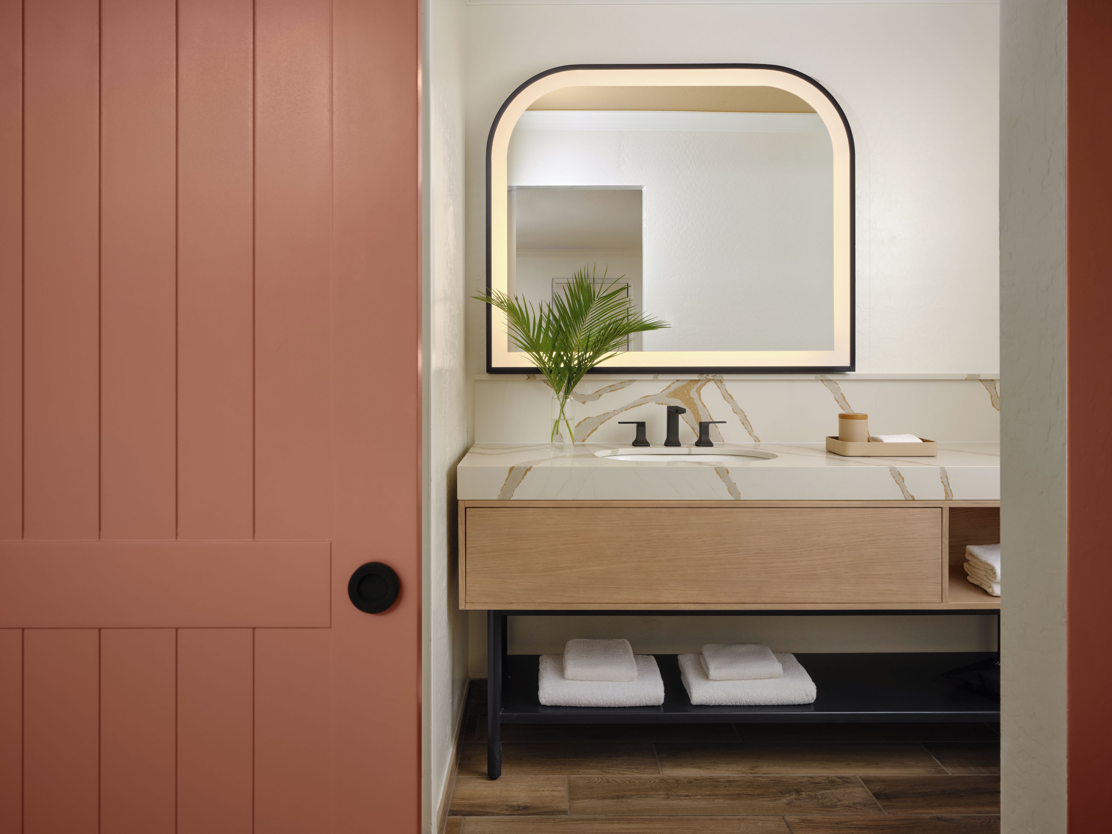 Un bagno con una porta in legno, un lavandino bianco con piano in marmo, un rubinetto nero, uno specchio con cornice nera, una pianta in vaso e due asciugamani su uno scaffale.