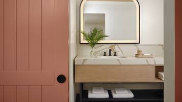 Un bagno con una porta in legno, un lavandino bianco con piano in marmo, un rubinetto nero, uno specchio con cornice nera, una pianta in vaso e due asciugamani su uno scaffale.
