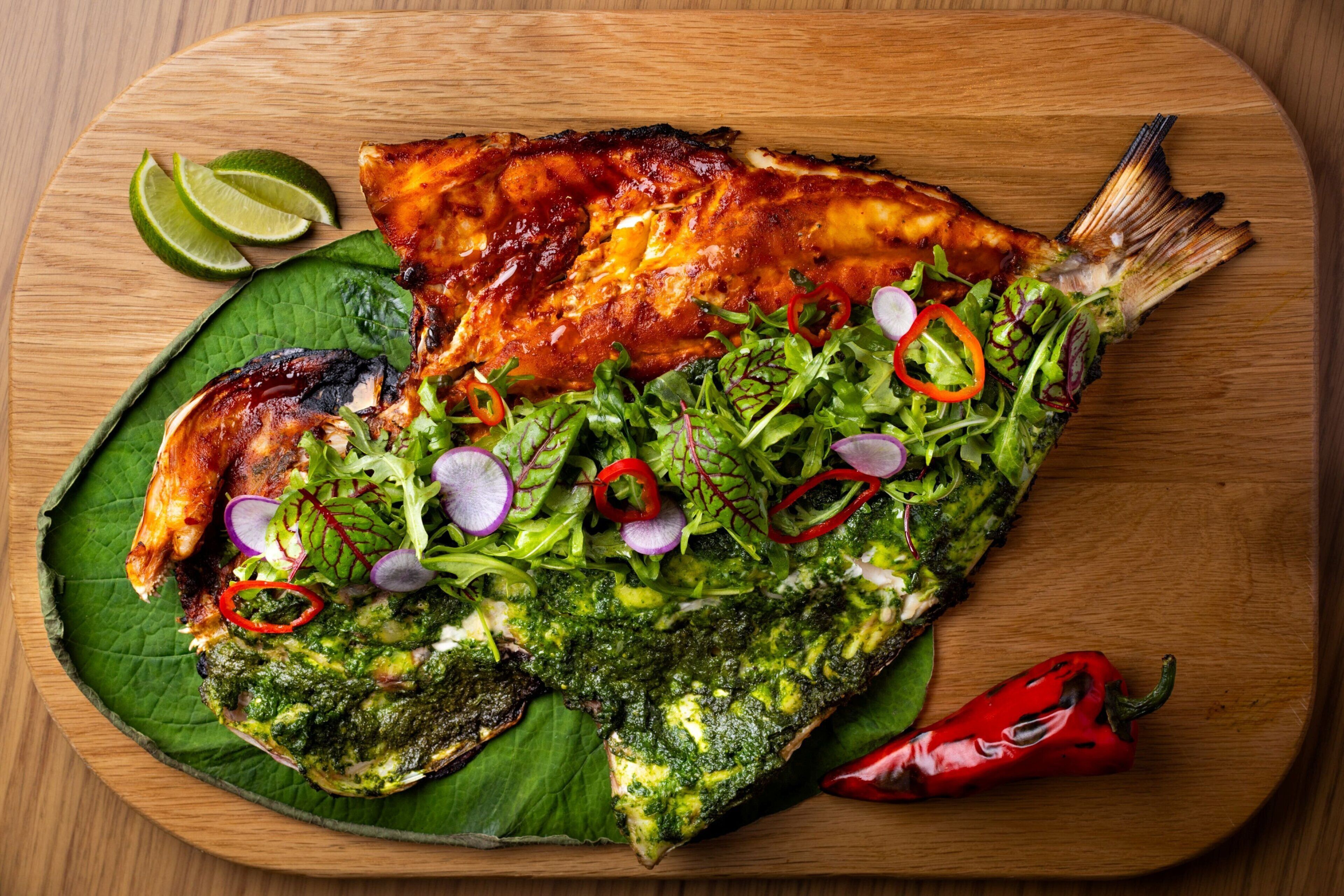 A grilled fish dish with greens and lime on a wooden board.