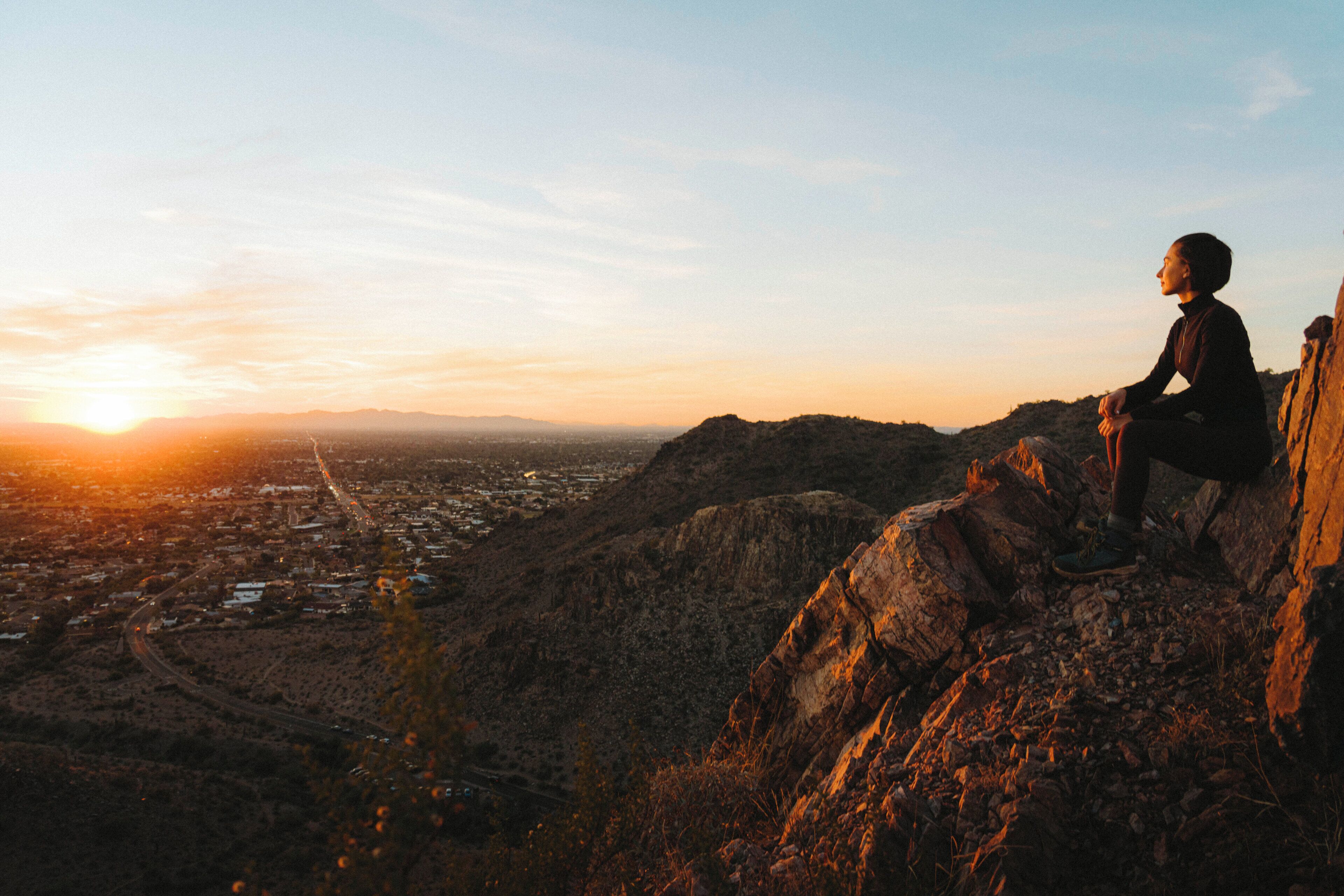 A person sitting on a rocky outcrop overlooking a cityscape during sunset.