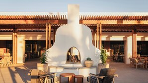 Un área de estar al aire libre con sillas y mesas de madera, una chimenea central y un edificio con techo de tejas al fondo.