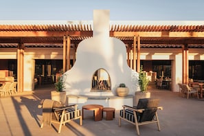 Een terras met houten stoelen en tafels, een centraal element met vuur en een gebouw met pannendak op de achtergrond.