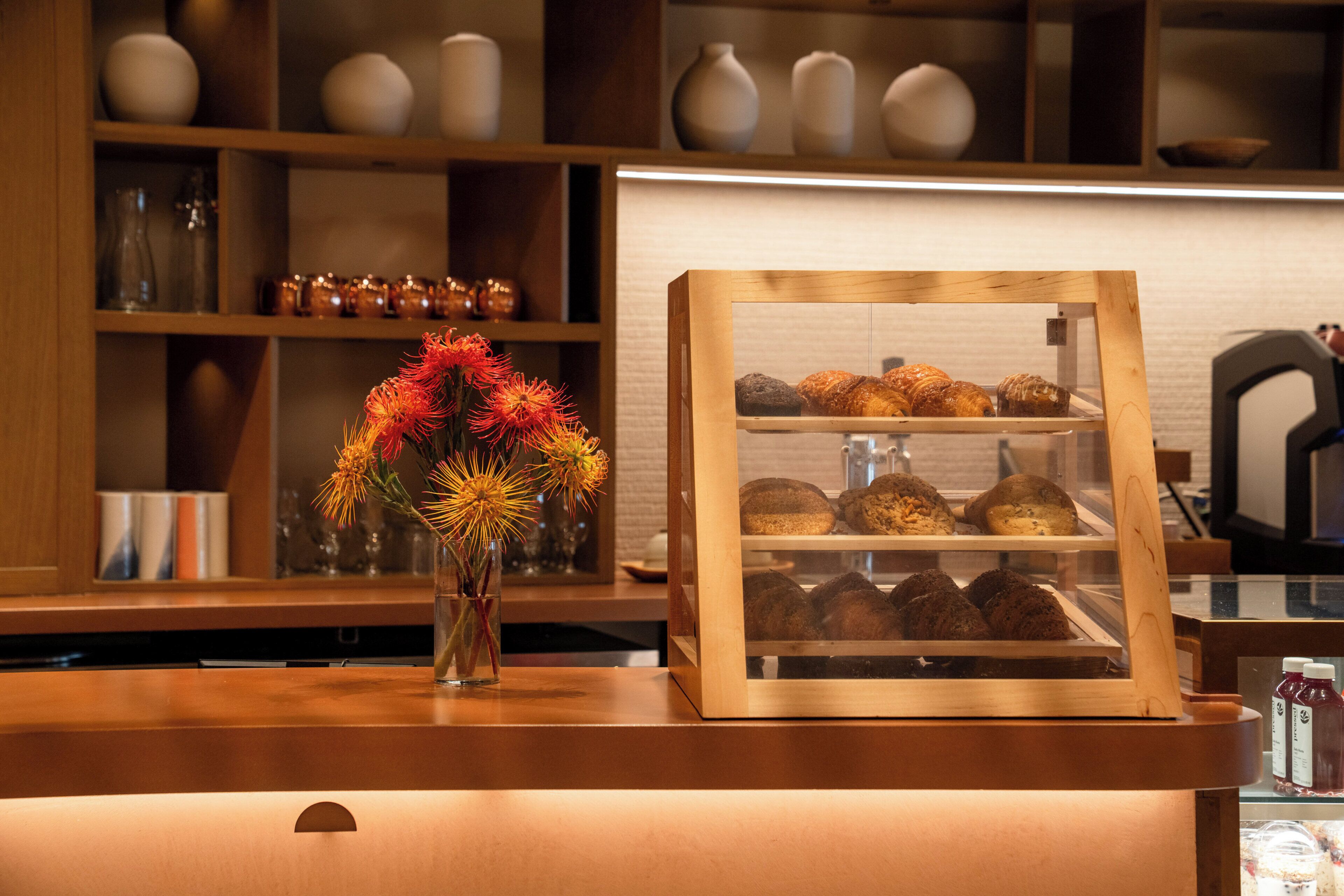 Una vitrina de pastelería con pasteles, un jarrón con flores y estantes con varios objetos al fondo.