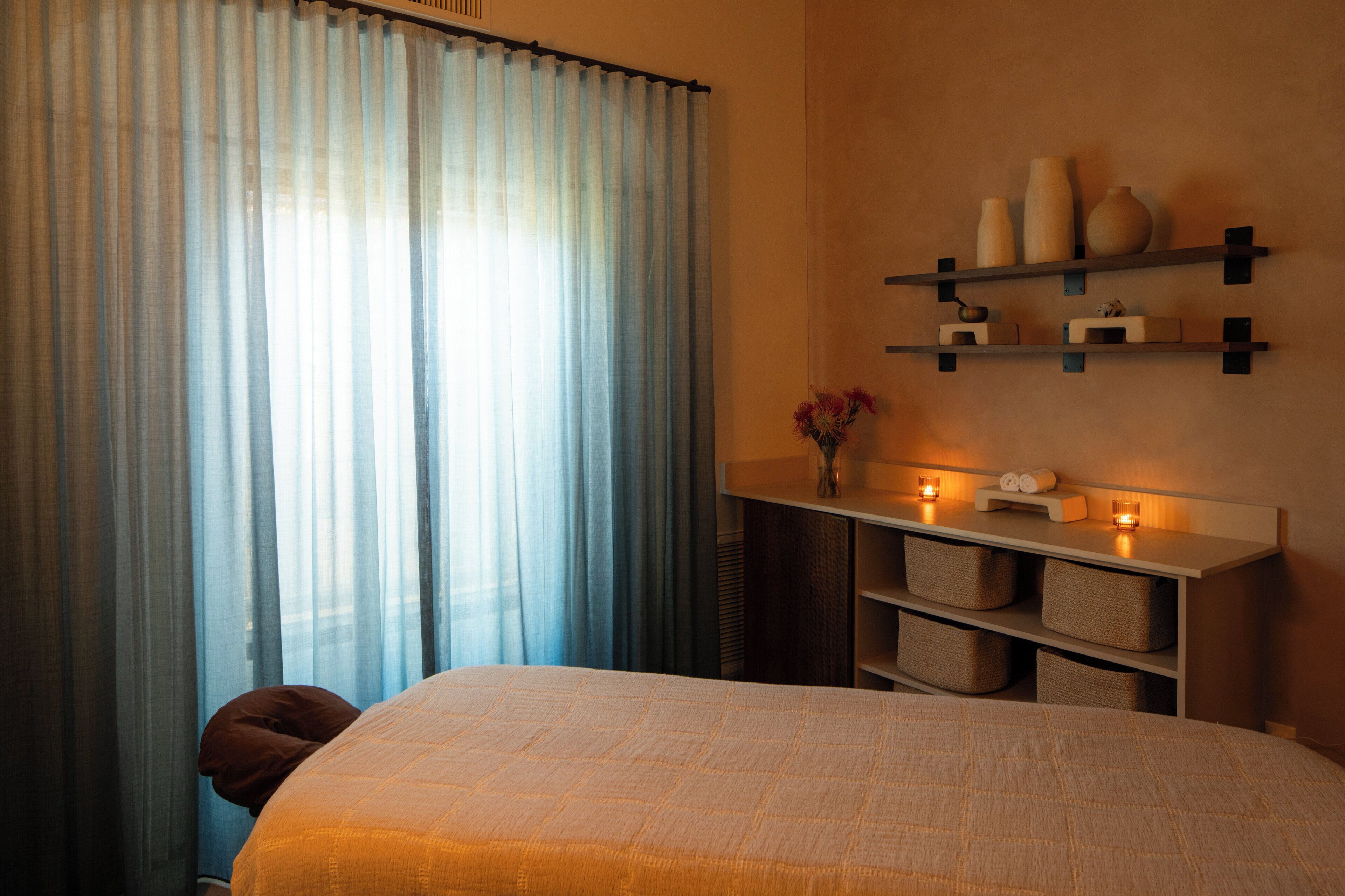 A bedroom with a bed, a nightstand, a wall-mounted shelf with decorative items, and sheer curtains.
