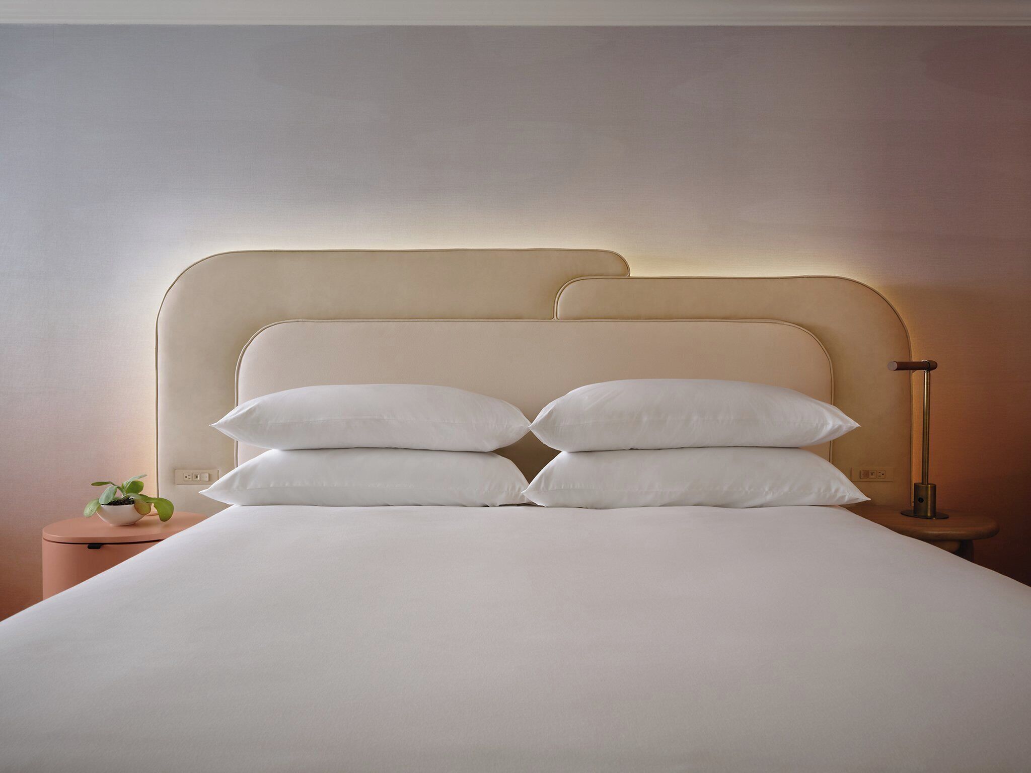 Una cama bien hecha con sábanas y almohadas blancas, una mesita de noche con una planta y una lámpara de pie.