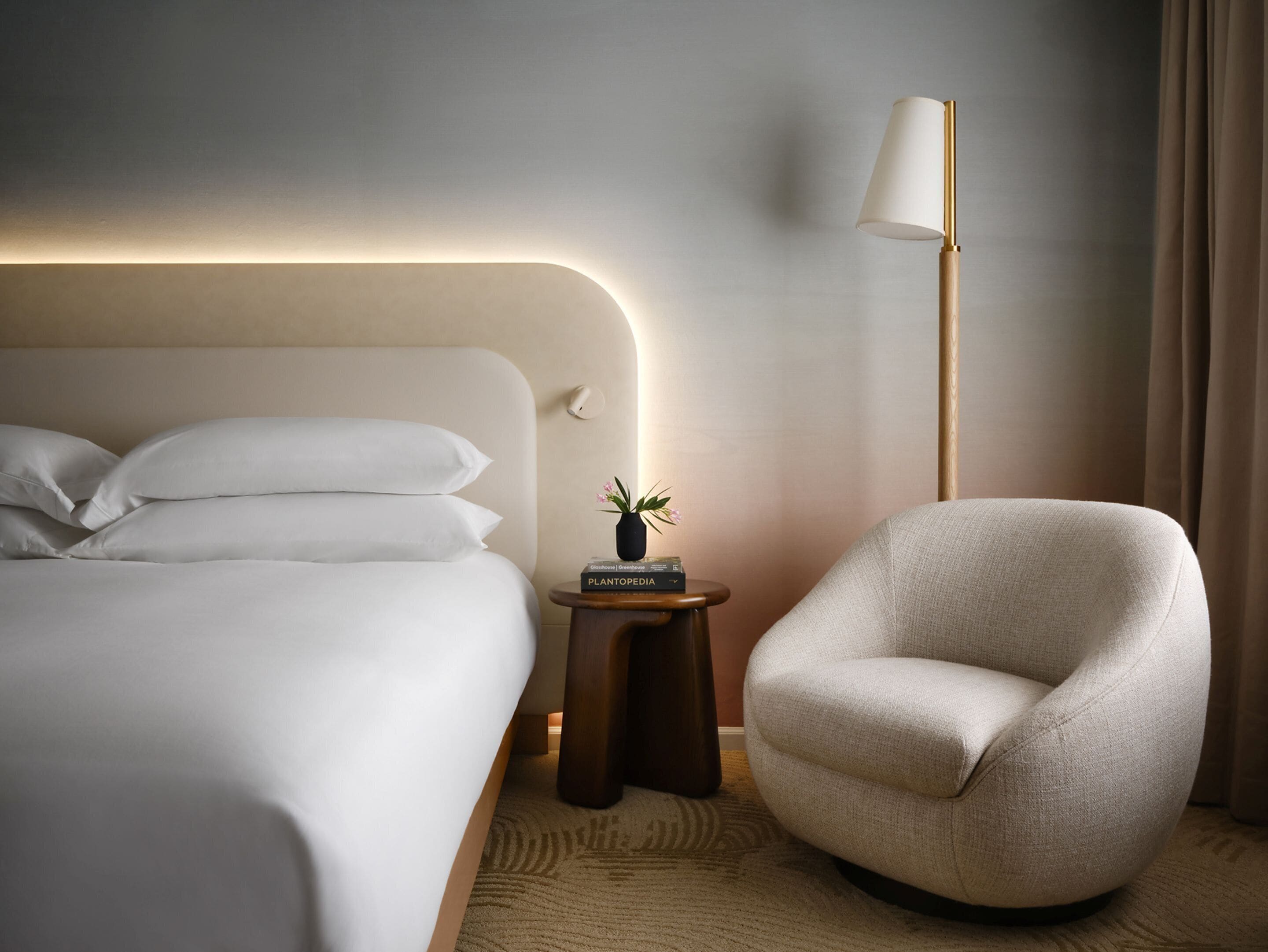 A modern hotel room with a neatly made bed, a small side table with a lamp, and a comfortable armchair.