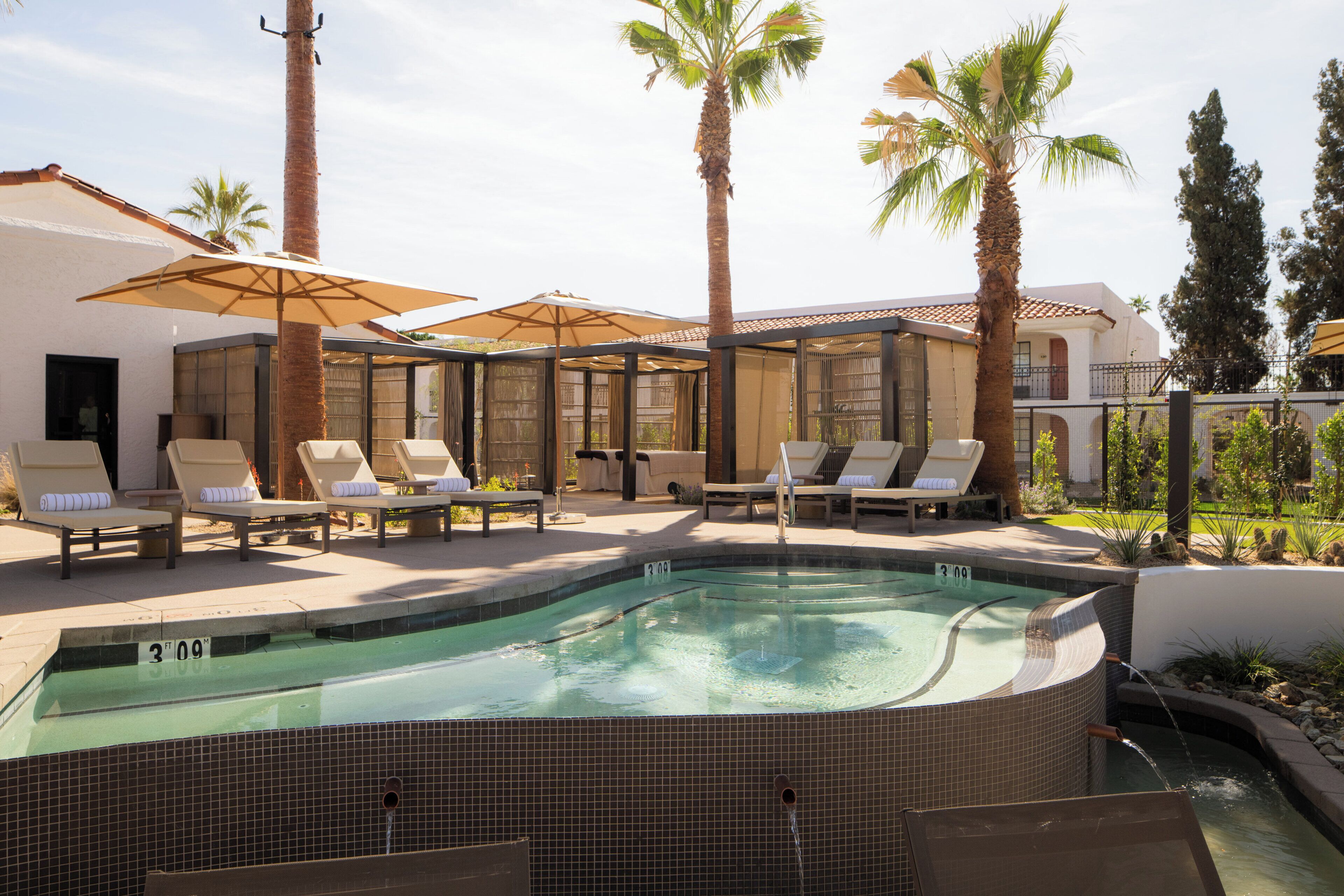 A swimming pool with lounge chairs and umbrellas, surrounded by palm trees and a building with a tiled roof.