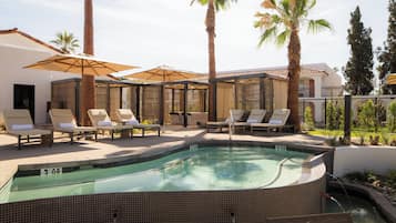 A swimming pool with lounge chairs and umbrellas, surrounded by palm trees and a building with a tiled roof.