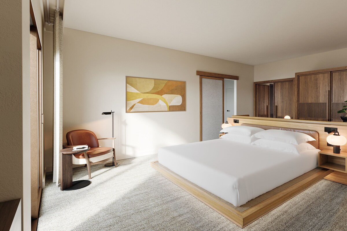 Ein Hotelzimmer mit Bett, Sessel, Schreibtisch, Schreibtischlampe, Nachttisch, Spiegel und einem Wandbild.