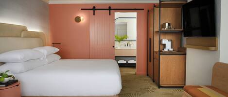 Habitación de hotel con una cama grande, un televisor montado en la pared, un pequeño rincón de descanso y un baño visible a través de una puerta abierta.