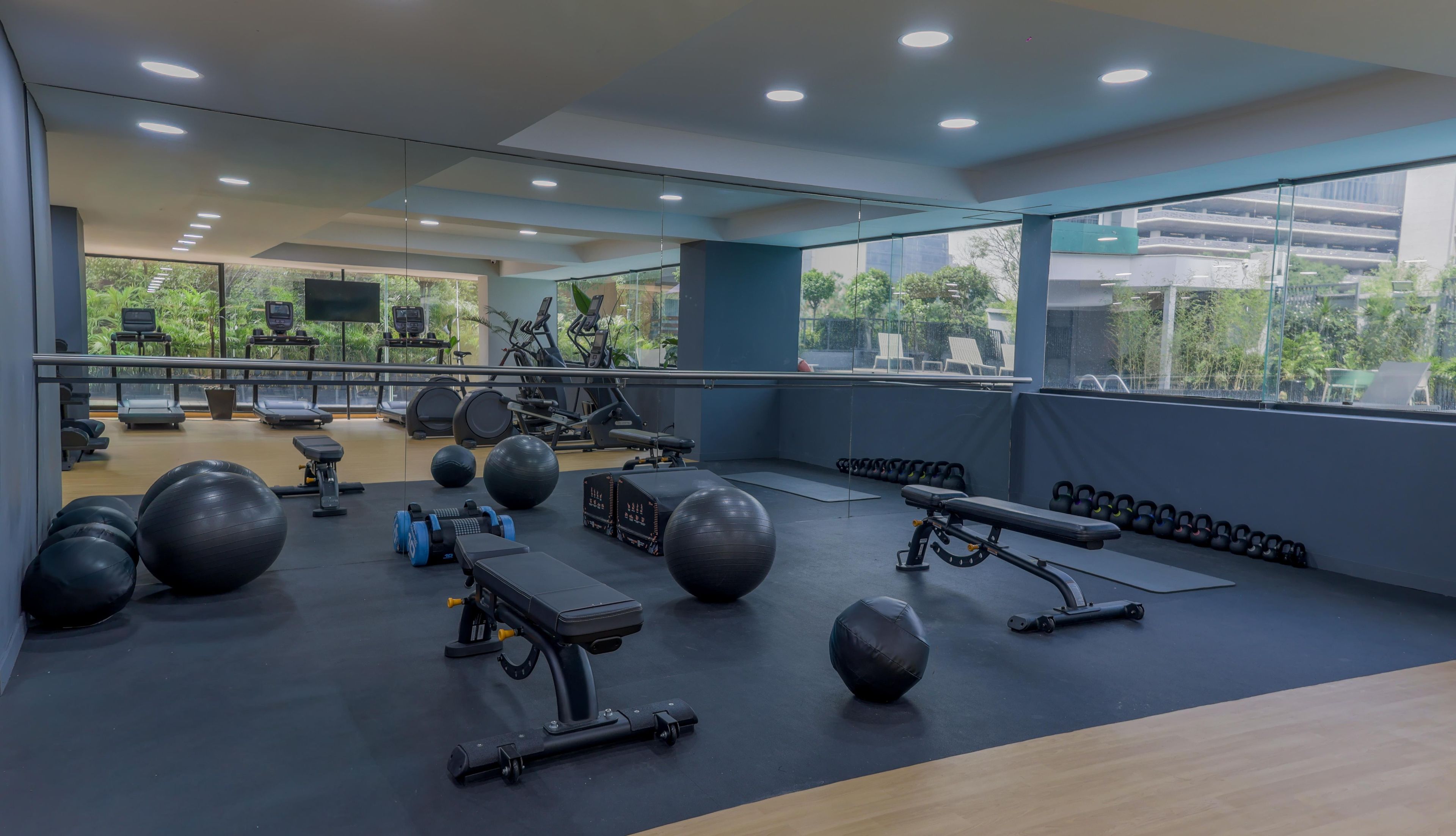 Un gimnasio con equipo de ejercicio, espejos y amplias ventanas con vistas a zonas verdes.