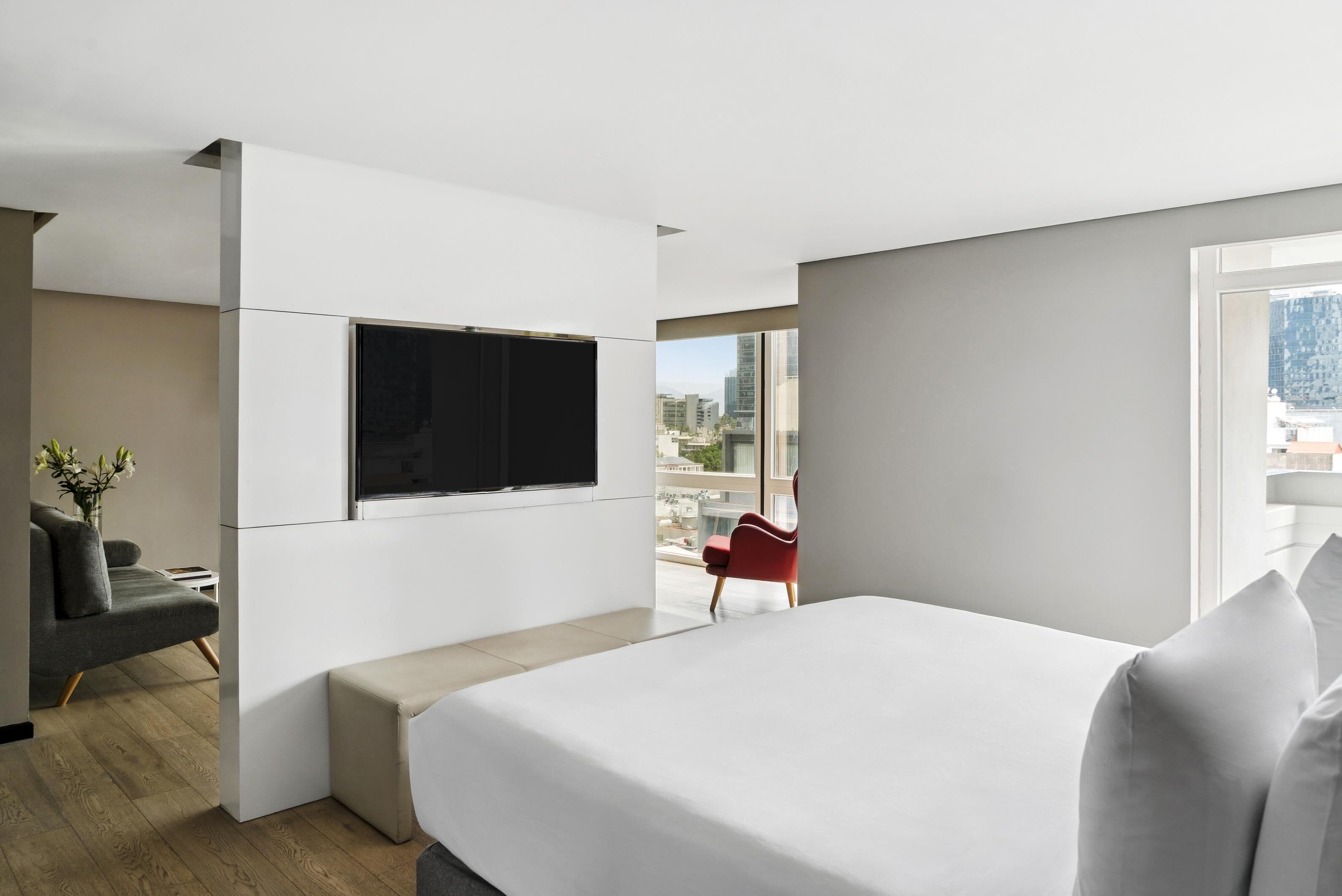 Una habitación de hotel moderna con una cama grande, un televisor de pantalla plana montado en la pared, una silla roja y vistas a la ciudad a través de amplias ventanas.