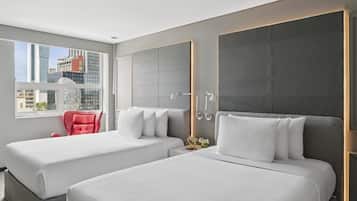 A modern hotel room with two beds, a red chair, a large window with a city view, and a wall-mounted light fixture.
