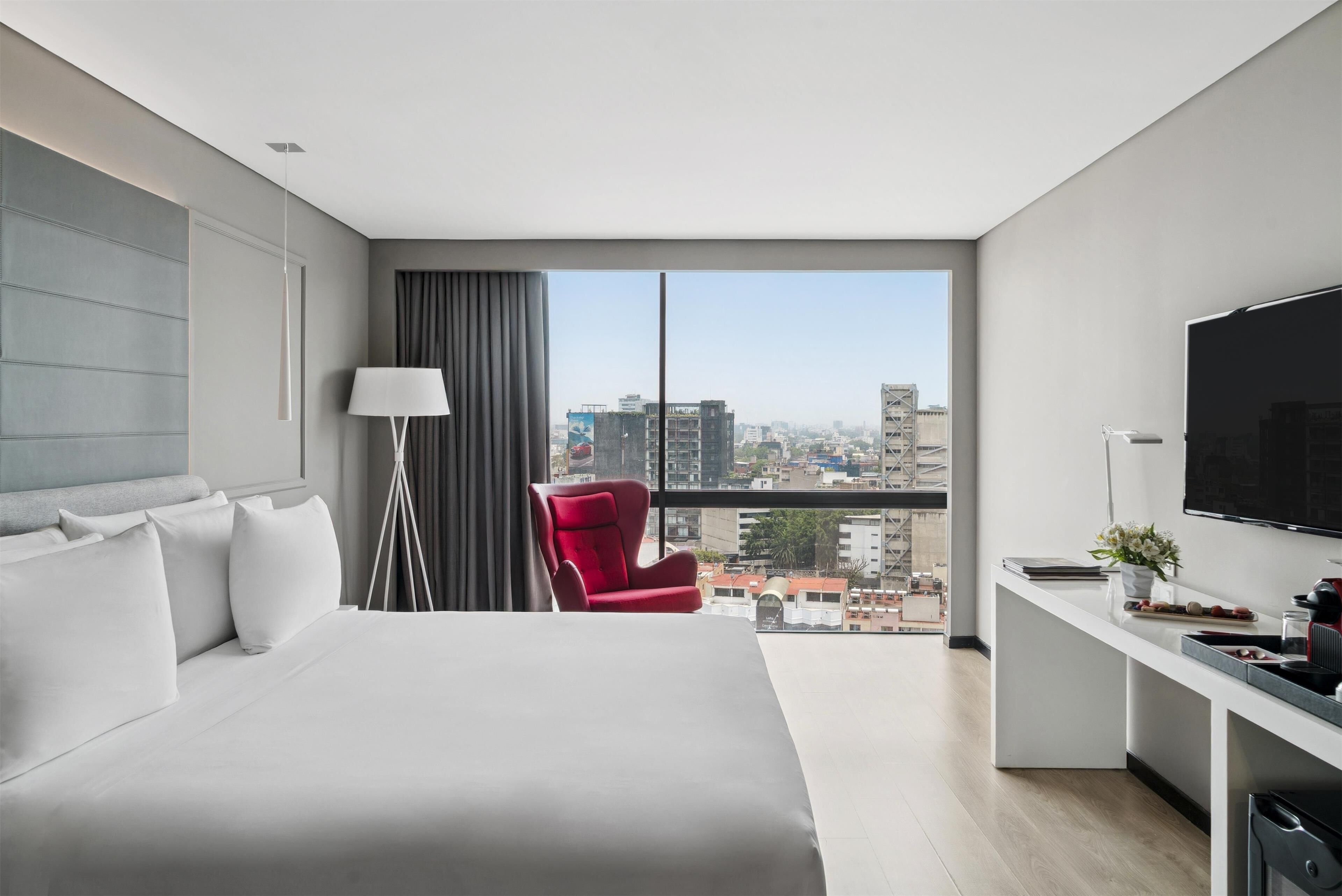 Une chambre d’hôtel moderne dotée d’un grand lit, d’un bureau avec un téléviseur à écran plat, d’un fauteuil rouge et offrant une vue sur la ville grâce à de grandes fenêtres.