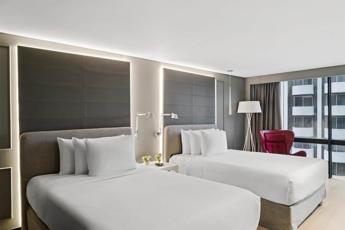 Ett hotellrum med två sängar, en röd stol, en lampa och utsikt över staden.