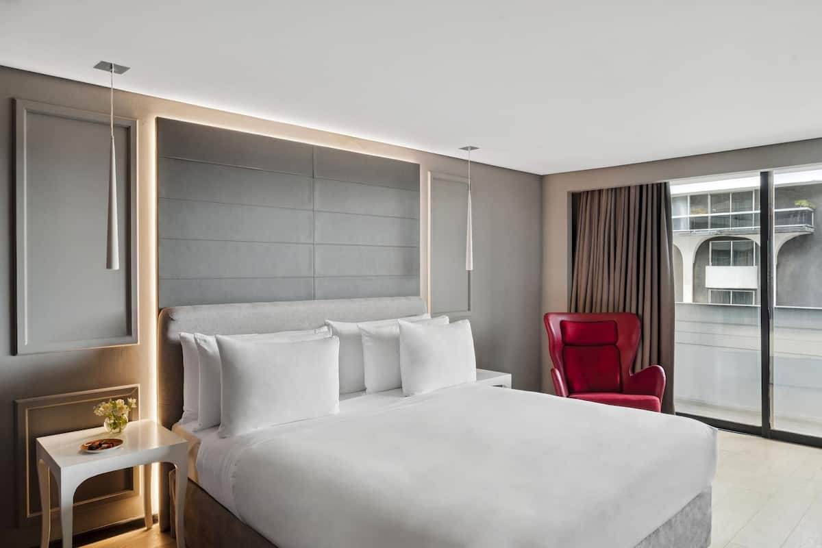 Ett modernt hotellrum med en stor säng, en röd fåtölj, ett litet bord med en vas med blommor och ett stort fönster med utsikt över staden.