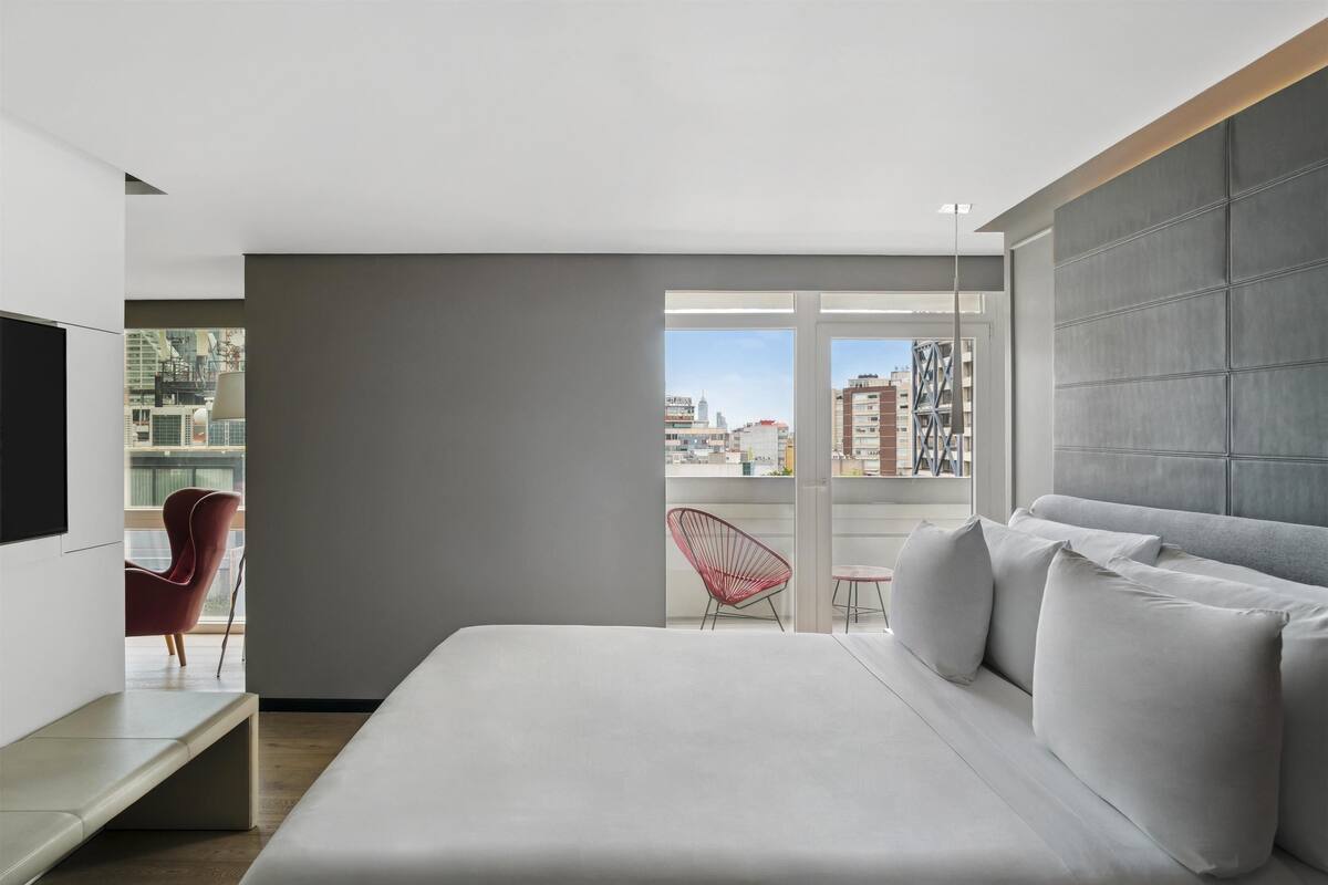Ett modernt hotellrum med en stor säng, en TV, en röd fåtölj och en balkong med utsikt över staden.