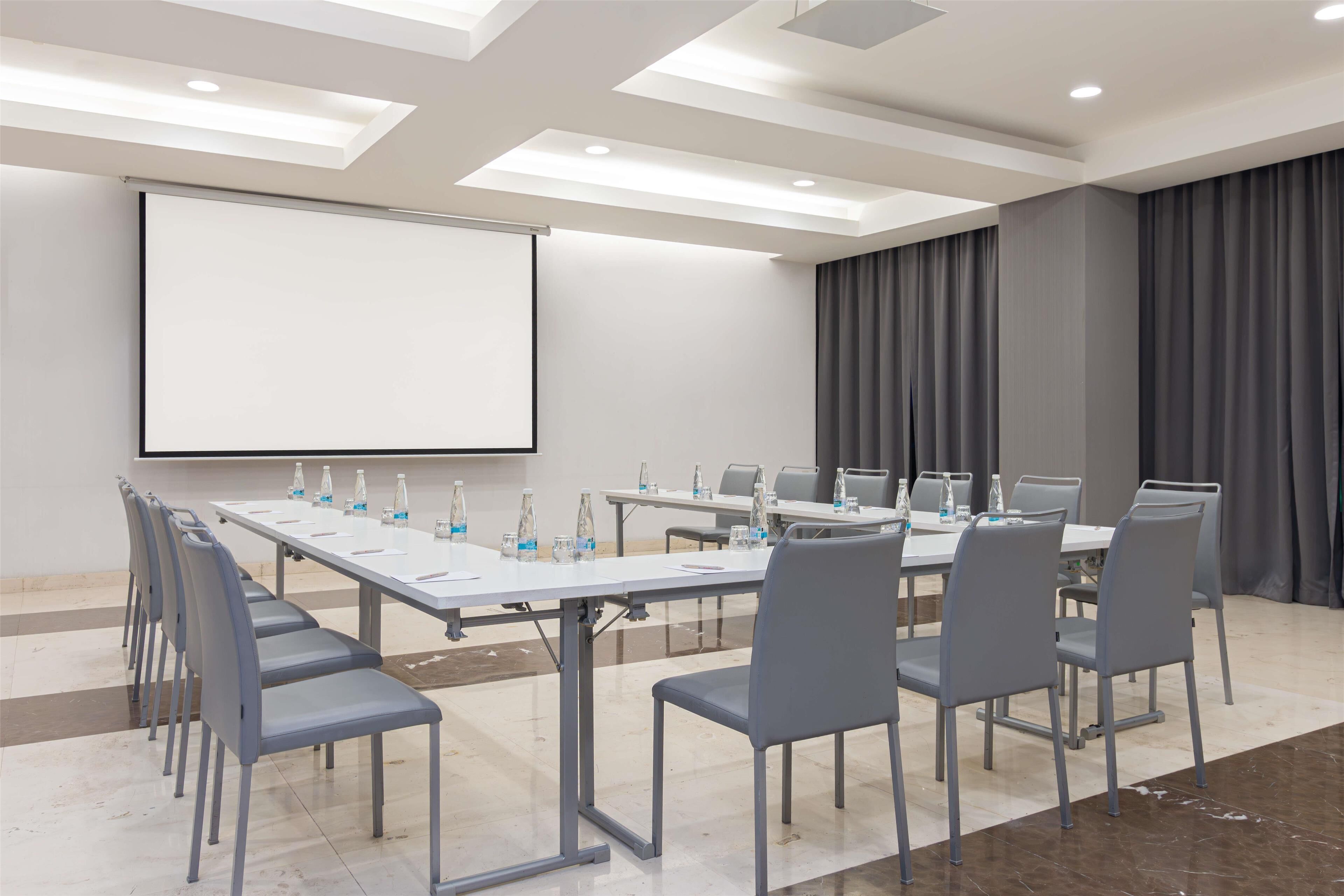A conference room with a long table, chairs, a projector screen, and a large window with curtains.