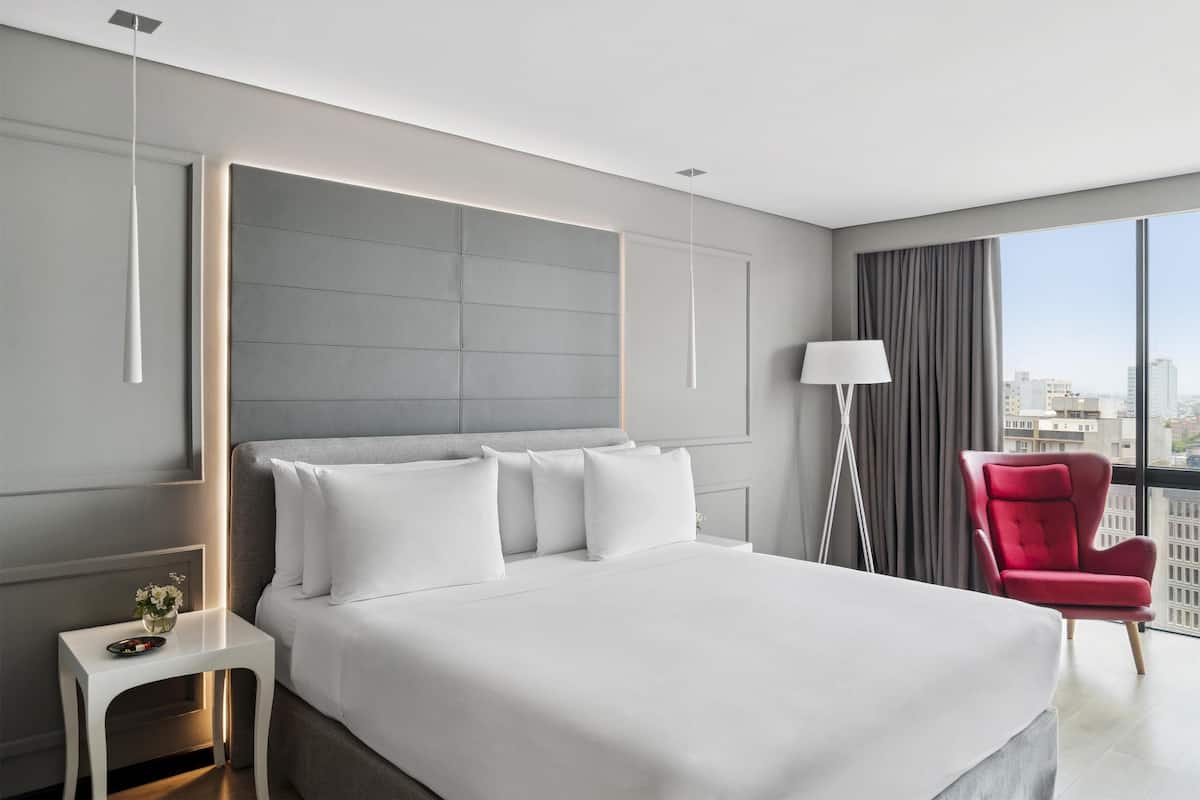 Ett modernt hotellrum med en stor säng, en röd fåtölj och utsikt över staden genom fönstret.