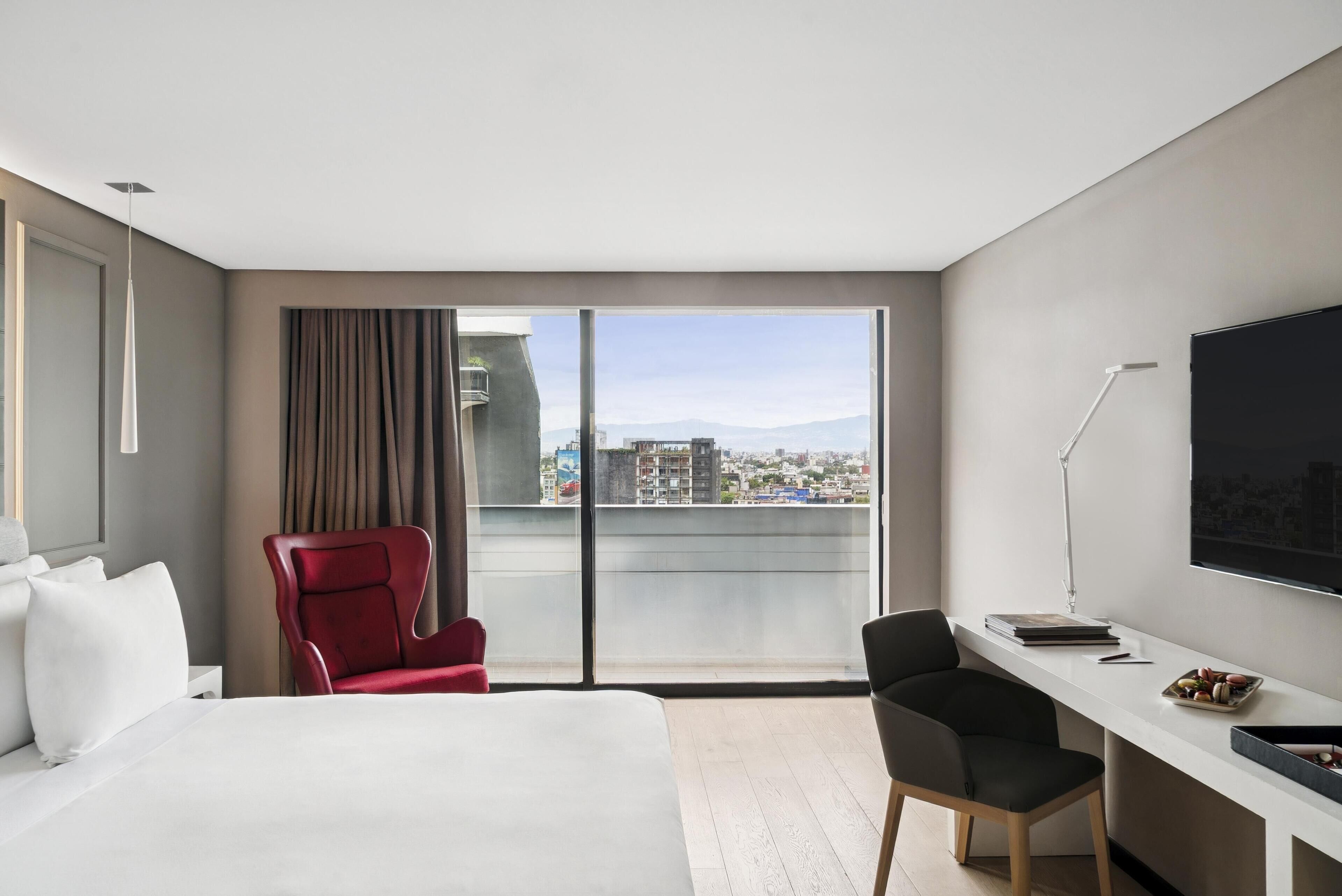 A modern hotel room with a large bed, a desk, a red chair, and a view of the city through large windows.