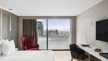 Una habitación de hotel moderna con una cama grande, un escritorio, una silla roja y vistas a la ciudad a través de amplios ventanales.