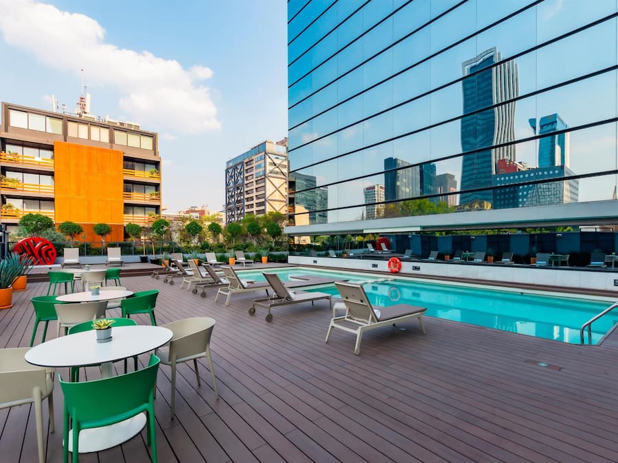 Uma área de piscina no terraço com espreguiçadeiras e mesas, cercada por plantas em vasos e vista para a cidade.