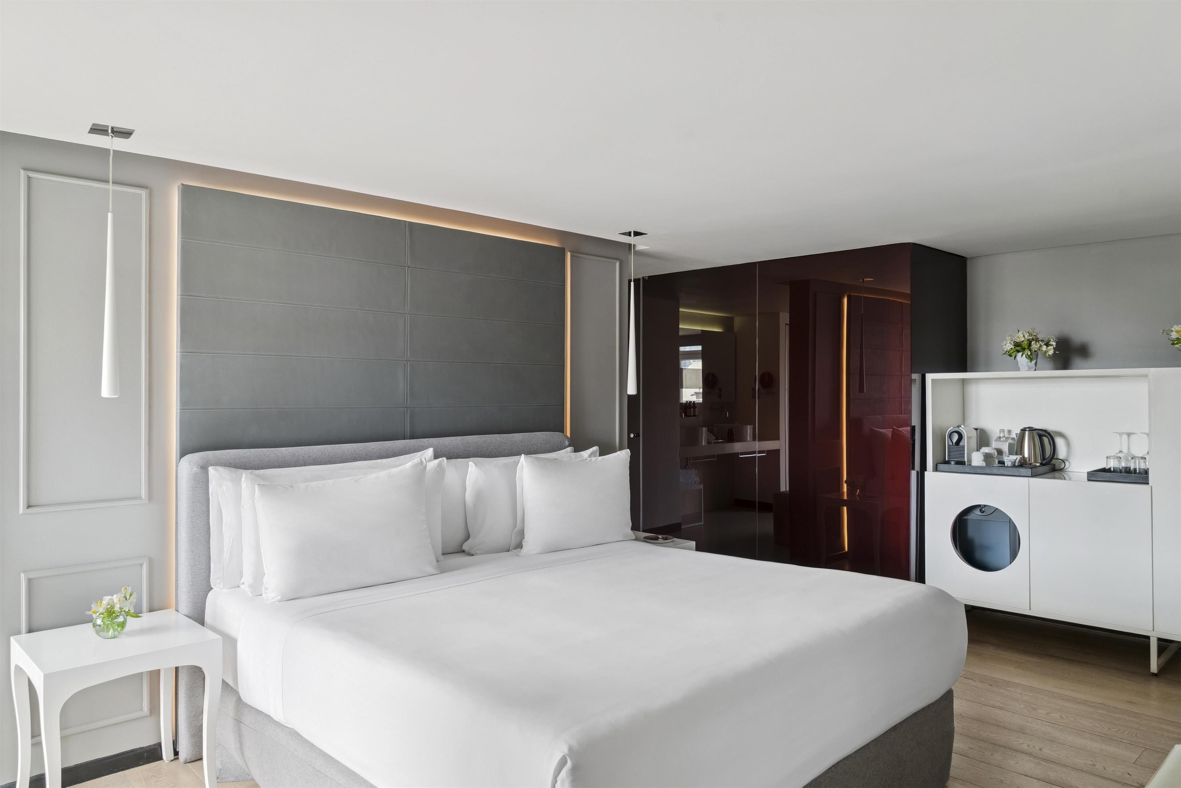 A modern hotel room with a large bed, a white nightstand, a wardrobe, and a small kitchenette area.