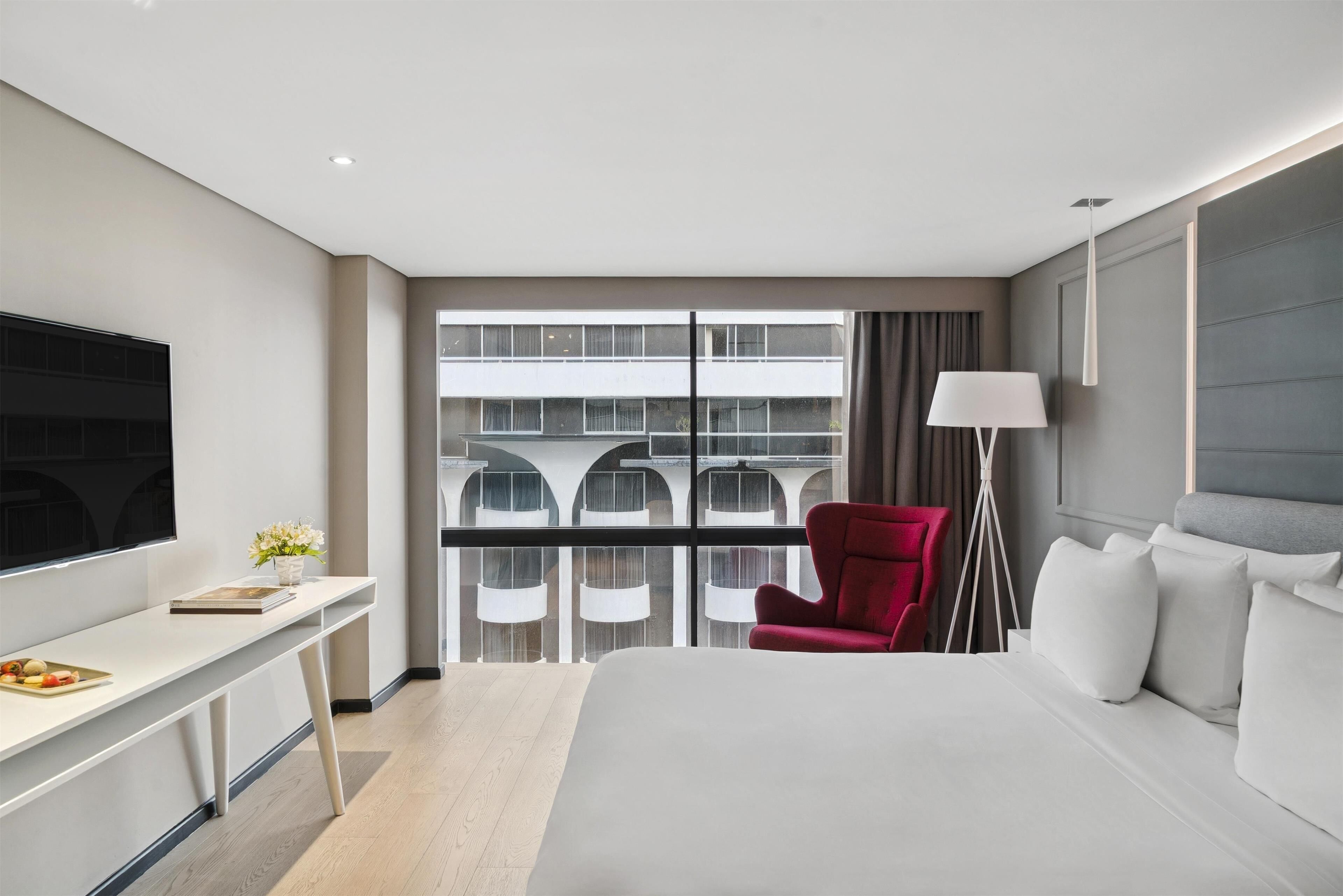 A modern hotel room with a large bed, a desk with a flat-screen TV, a red armchair, and a floor lamp.