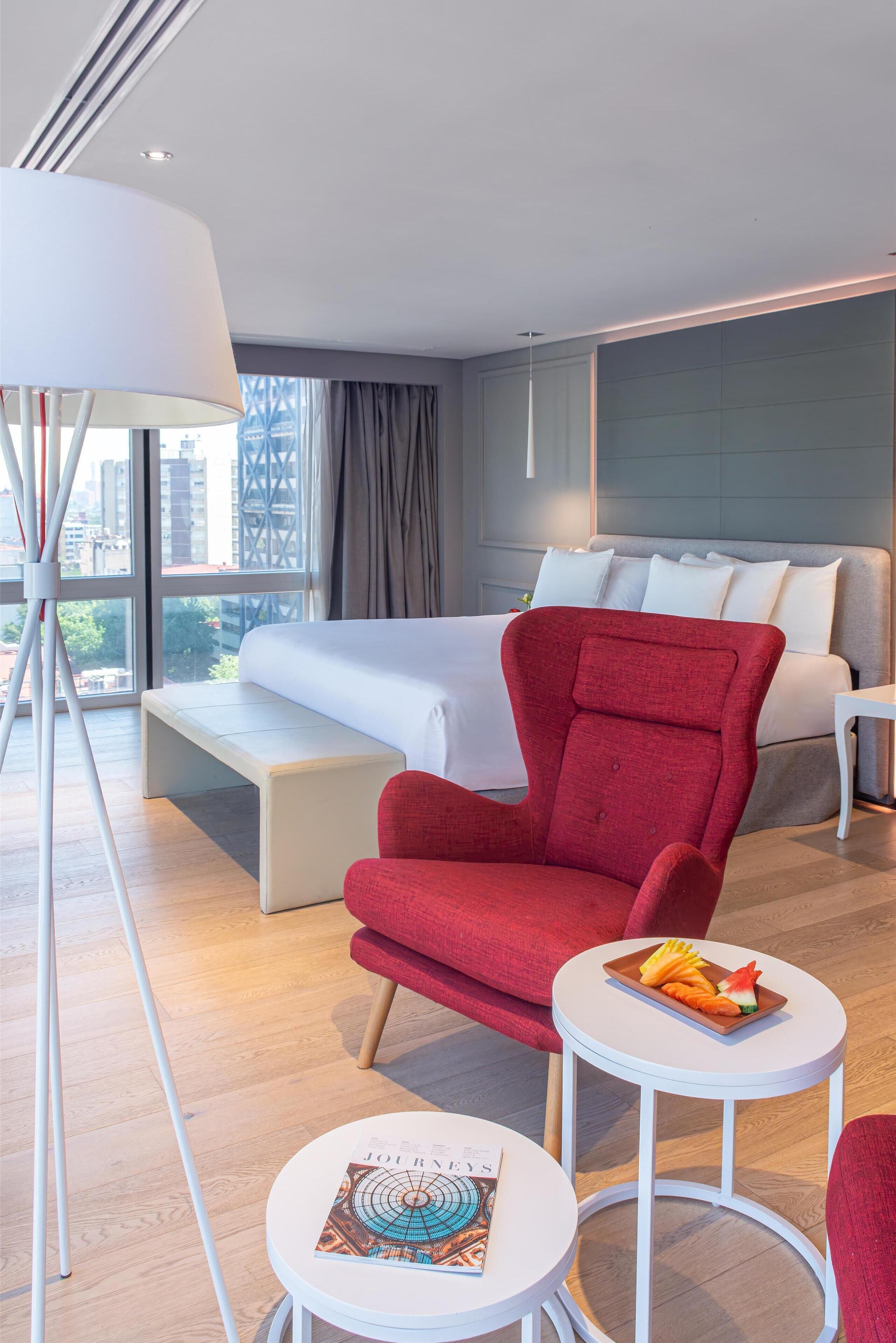 A hotel room with a bed, a red armchair, a small table with a fruit plate, and a magazine on the table.