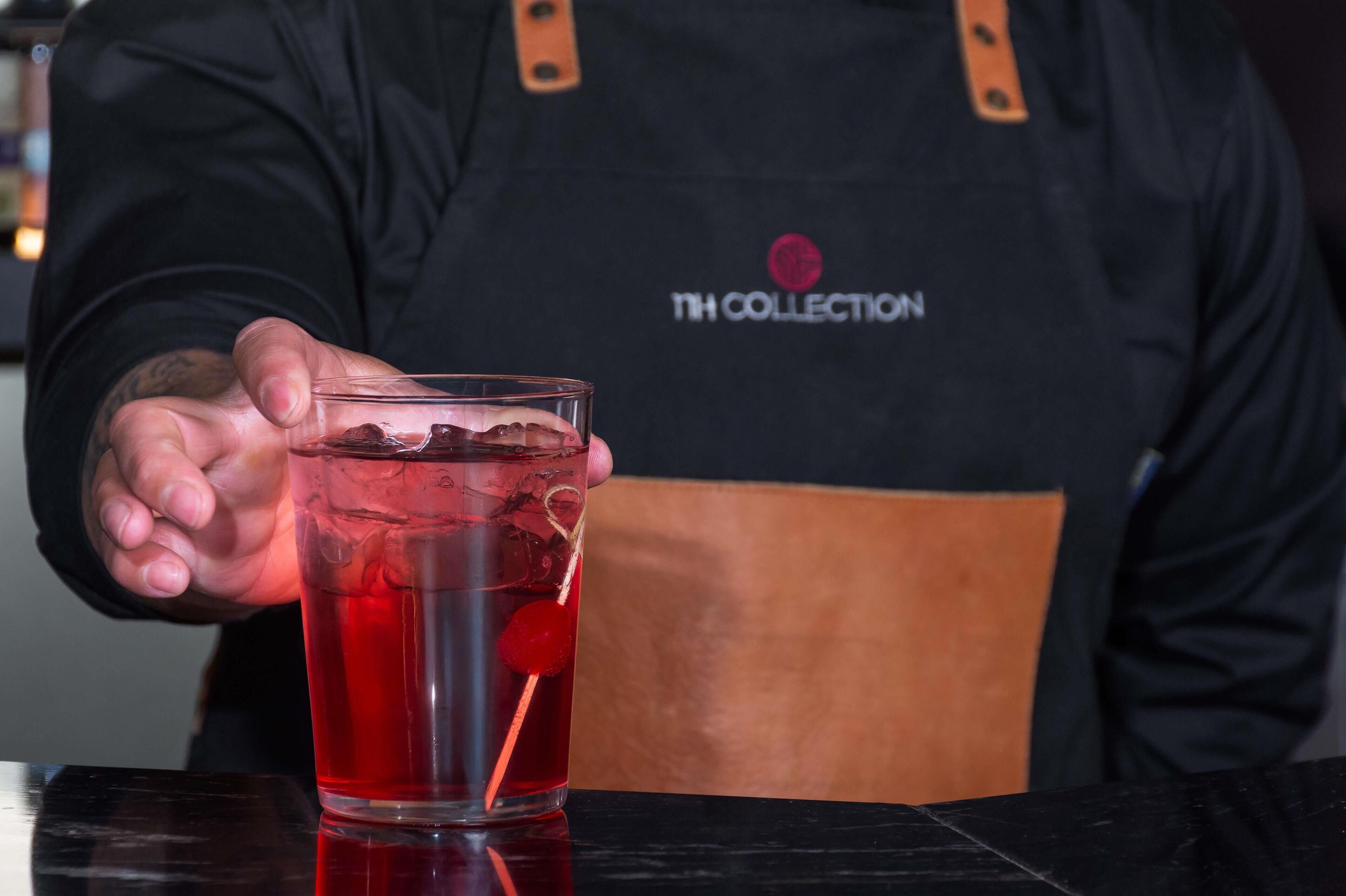 Eine Person in einer schwarzen Schürze mit einem Logo hält ein Glas mit einer roten Flüssigkeit und Eiswürfeln.