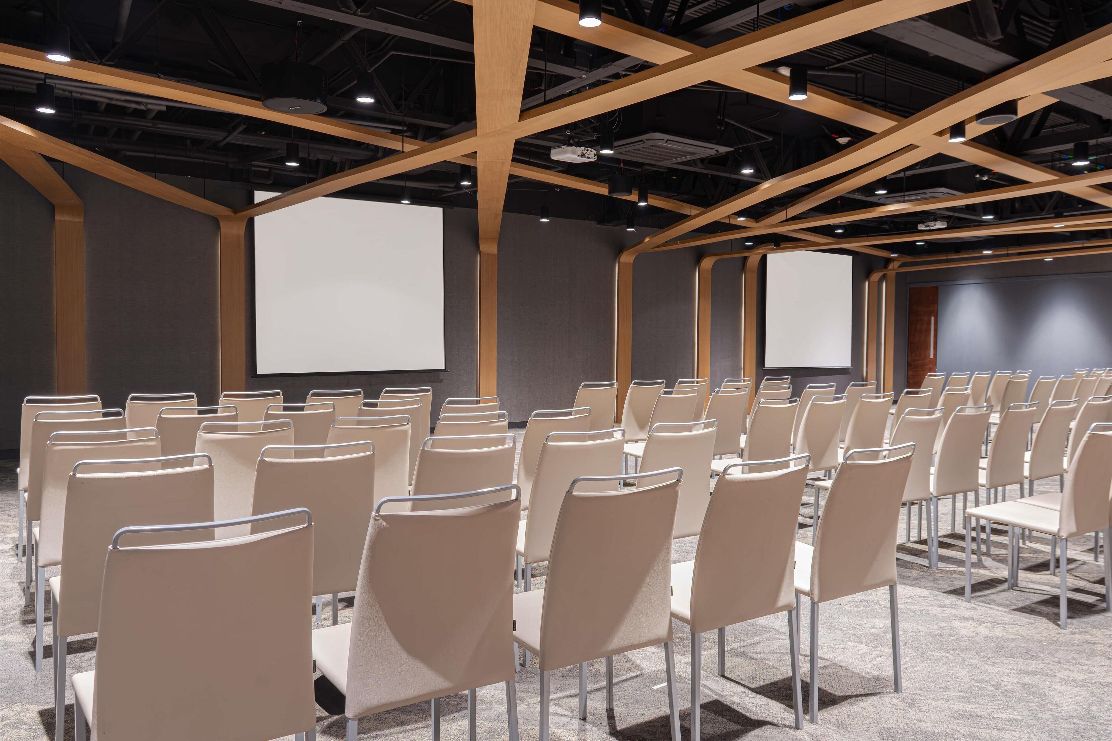 Un salón de conferencias con filas de sillas, una pantalla de proyector y una pared oscura.