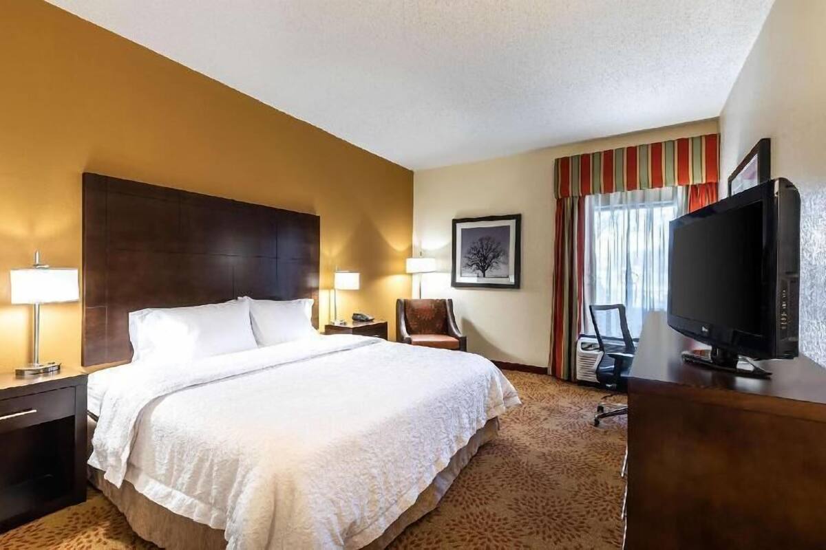 A hotel room with a large bed, two bedside lamps, a chair, a desk with a TV, and a framed picture on the wall.