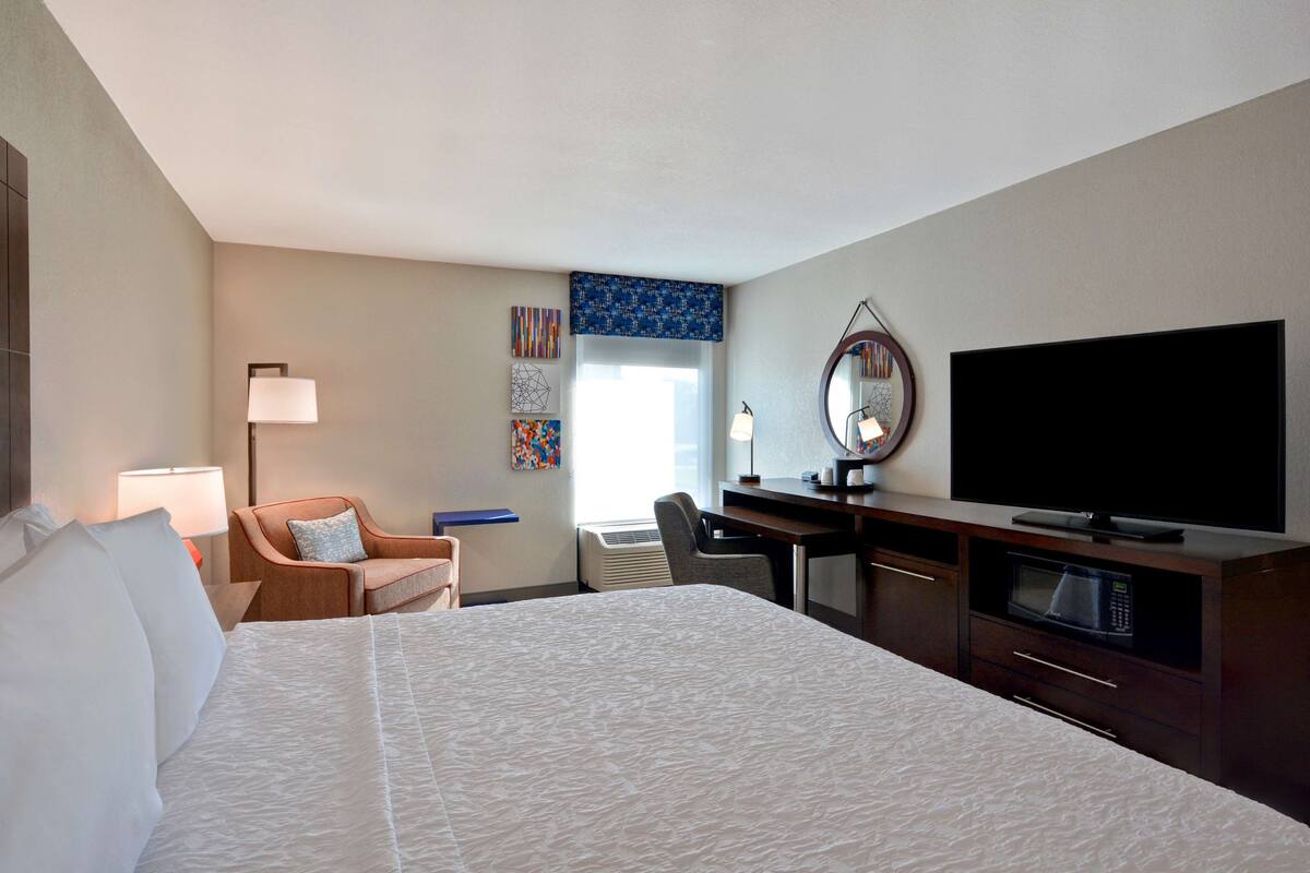 A hotel room with a large bed, a desk with a flat-screen TV, a chair, a lamp, and a microwave.