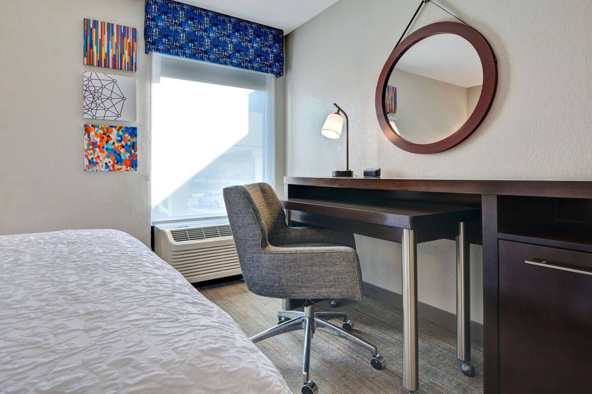 A hotel room with a bed, a desk with a chair, a round mirror, and artwork on the wall.
